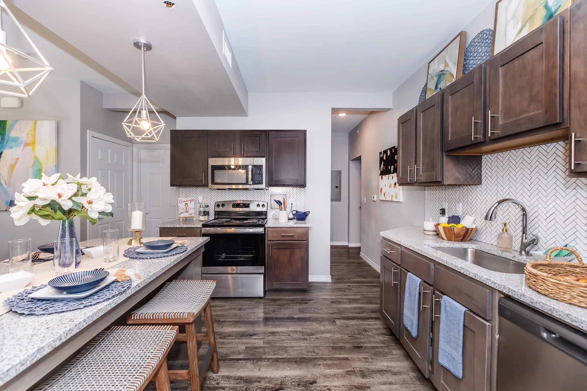 Modern kitchen featuring dark wooden cabinets, stainless steel appliances, and a granite countertop. A dining area with stylish tableware and decor complements the open layout. Pendant lights hang above the island, and stylish artwork adorns the walls, creating a warm and inviting atmosphere.