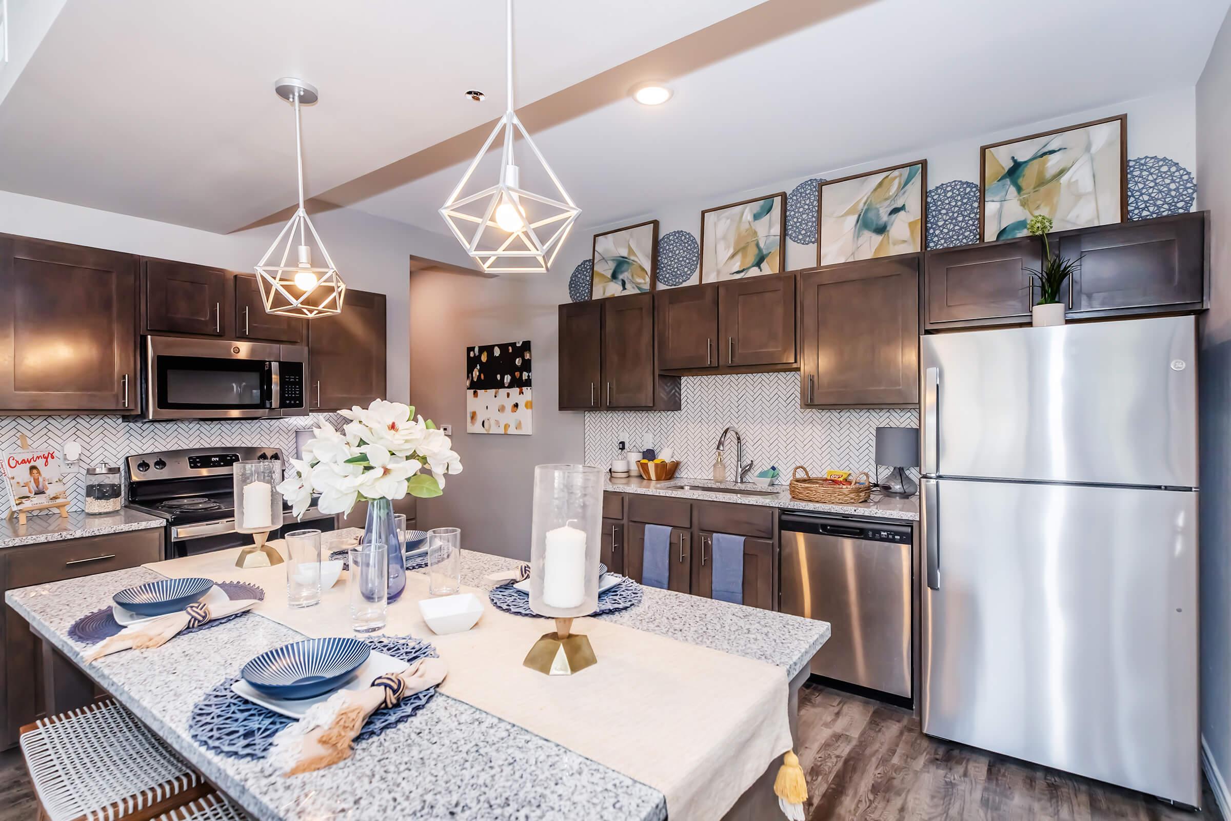 Modern kitchen featuring dark wooden cabinets, stainless steel appliances, and a marble countertop. A dining table is set with plates and glasses, decorated with flowers. The walls are adorned with framed artwork, and pendant lights hang overhead, creating a stylish and inviting atmosphere.