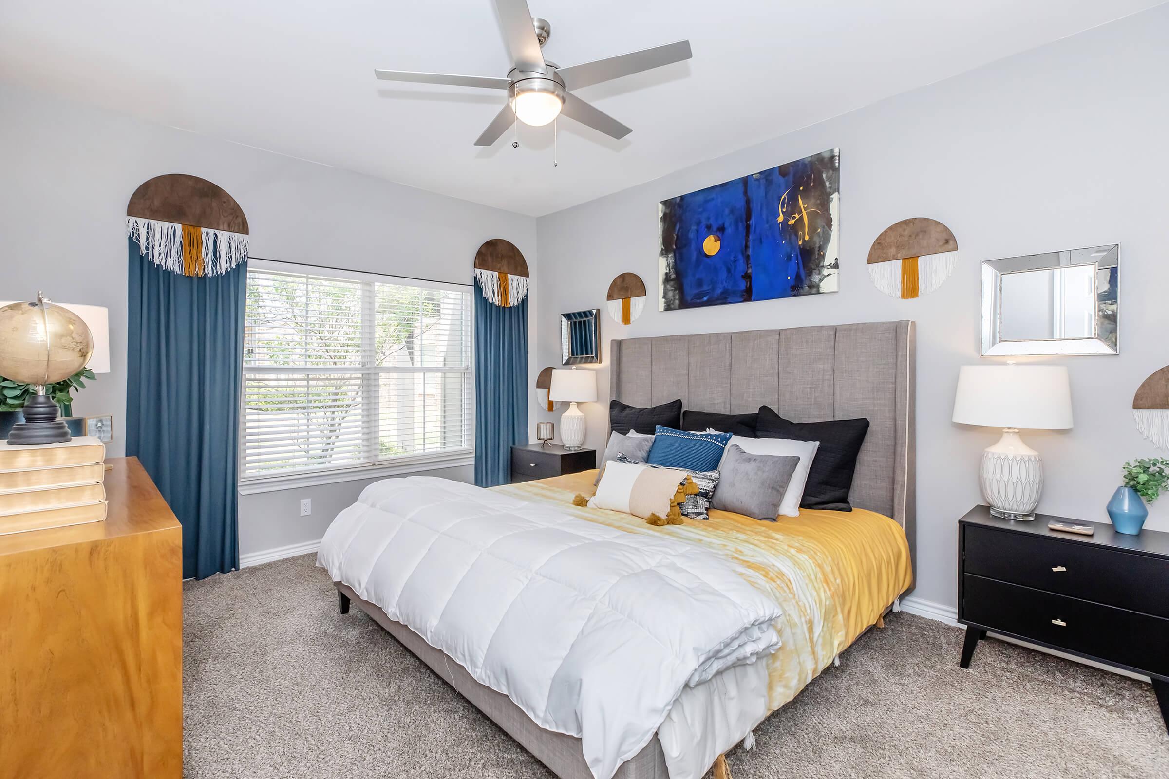 A stylish bedroom featuring a gray upholstered bed with yellow and blue bedding. There are two nightstands with lamps on either side, a window with blue curtains, and decorative wall art. The room has a soft, neutral color scheme with a cozy carpeted floor and modern ceiling fan.