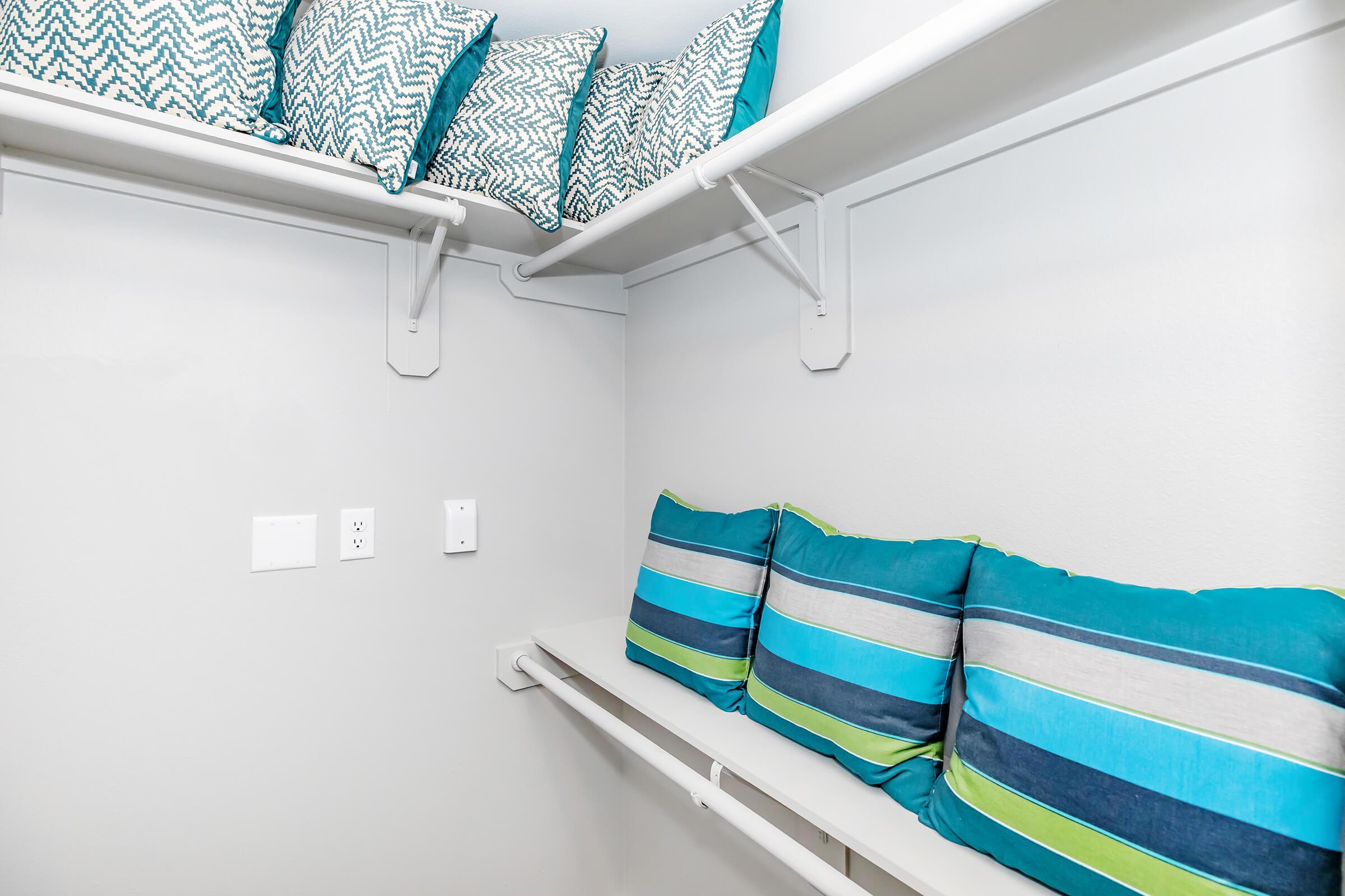 A closet featuring two shelves with decorative pillows in various shades of blue and green, and a built-in bench with striped pillows. The walls are painted gray, and there are two electrical outlets visible on the wall.