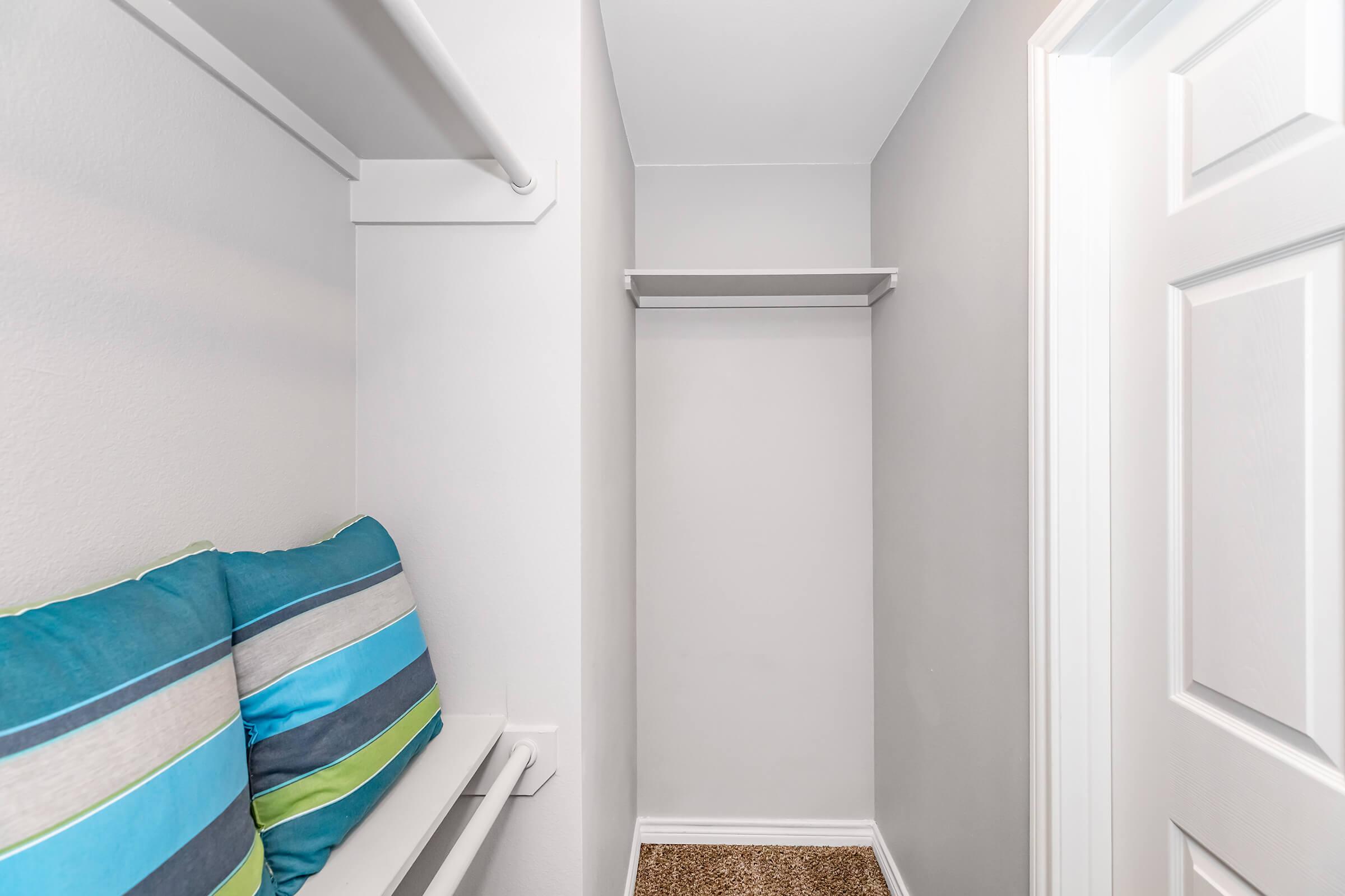 A small, well-lit closet with light gray walls. The closet features a shelf on the left and a small cushioned seating area with blue and green striped pillows. The floor is carpeted, and the door on the right is closed. The space appears organized and minimalist.