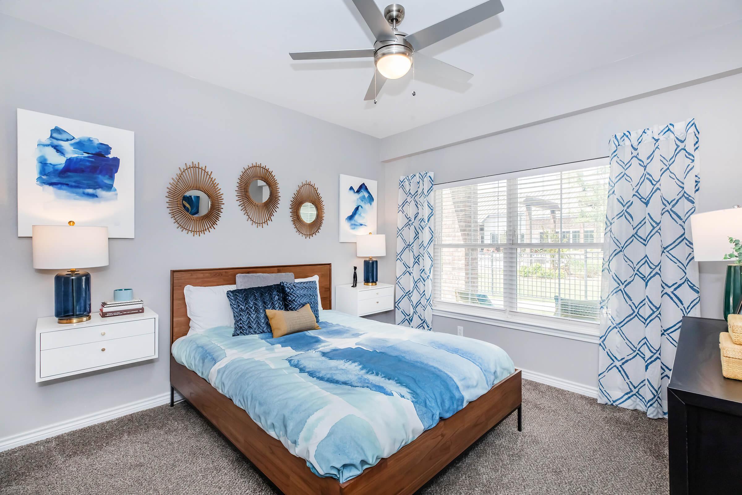 A modern bedroom featuring a queen-sized bed with a blue and white striped comforter, accented by decorative pillows. Framed artwork in shades of blue adorns the walls, complemented by circular mirrors. White nightstands with lamps flank the bed. Natural light streams in through a window with patterned curtains.