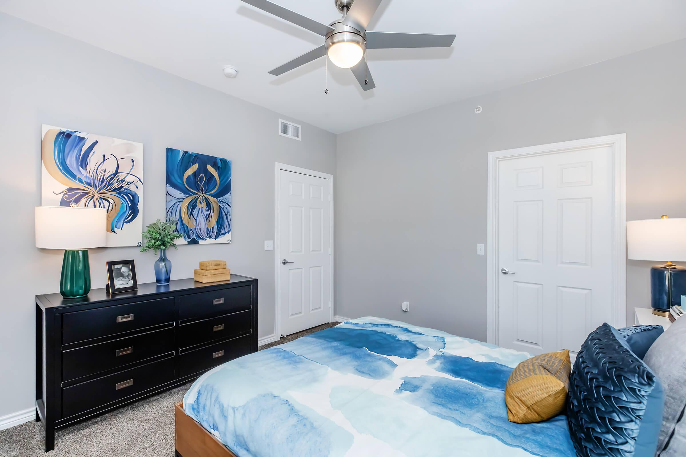 A cozy bedroom featuring a queen-sized bed with a blue and white abstract comforter, a modern dresser, and decorative artwork on the walls. A ceiling fan adds a contemporary touch, and there are two white doors leading to other areas. Soft lighting creates a warm ambiance.