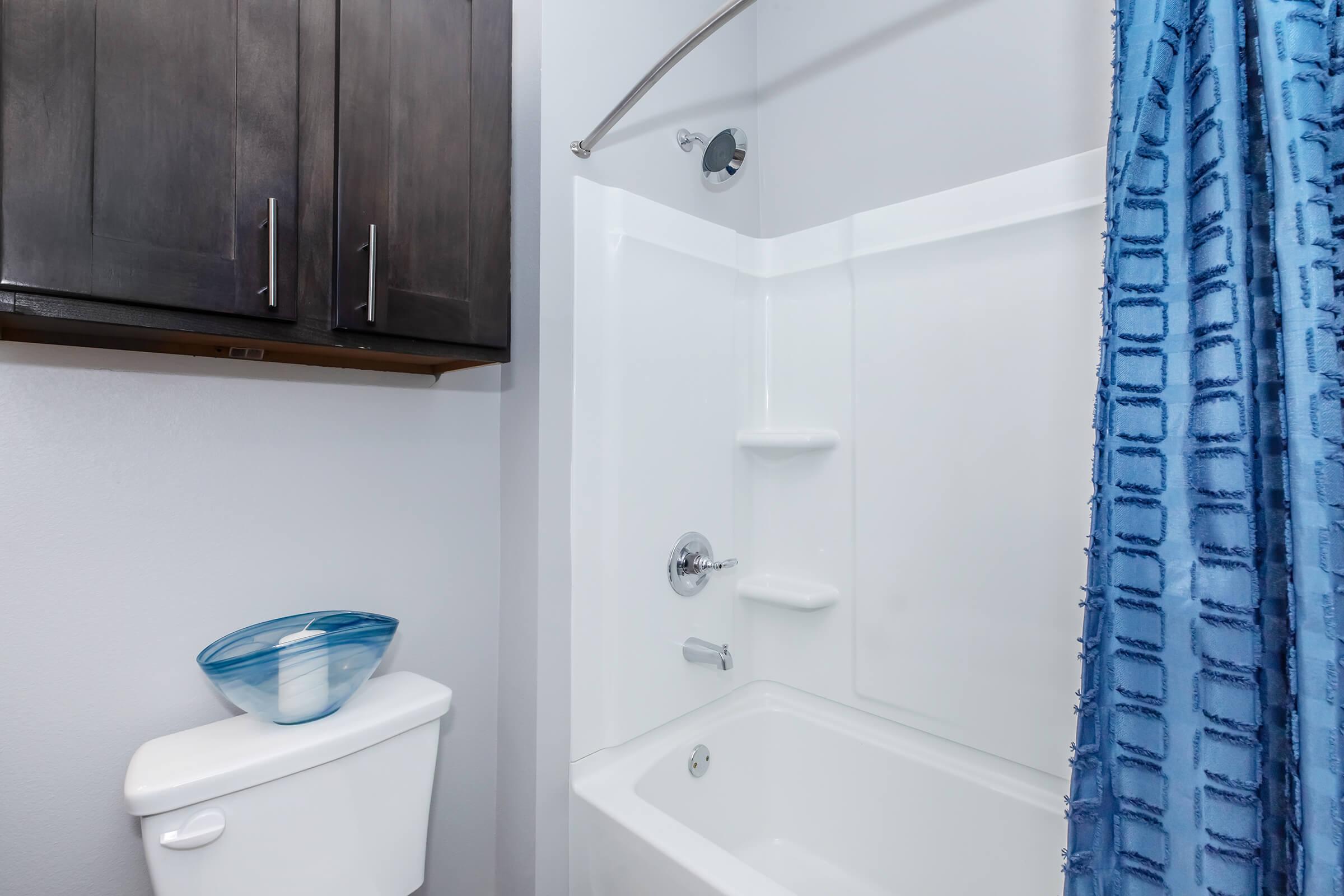A modern bathroom featuring a white bathtub and shower, a blue shower curtain with a textured pattern, a wall-mounted cabinet above, and a toilet nearby. The walls are painted light gray, creating a clean and contemporary look.