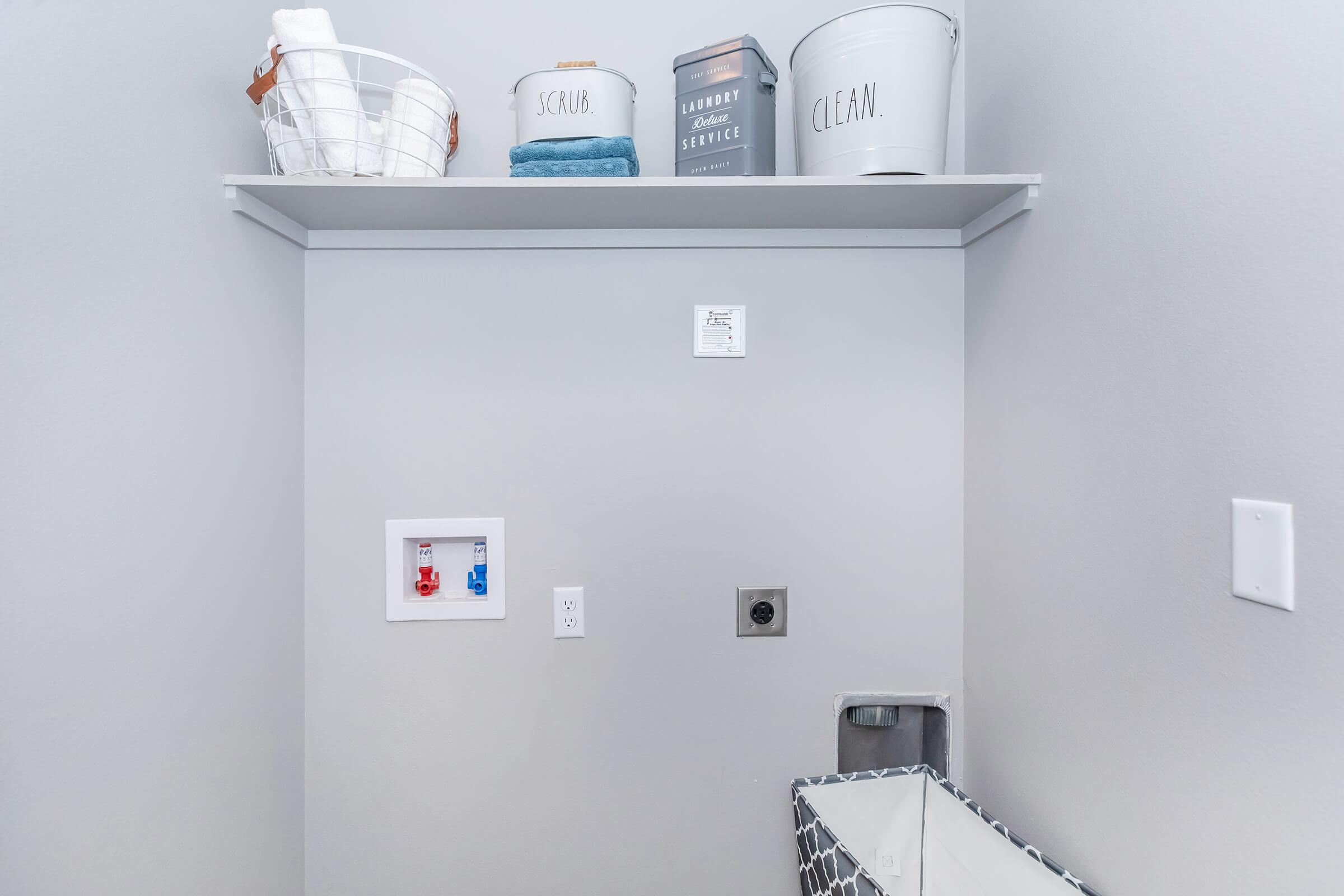 A laundry area featuring a shelf with neatly arranged items, including a basket, towels, and storage containers labeled 'SCRUBS' and 'CLEAN.' The wall has a small utility panel and an electrical outlet, with a laundry hamper placed on the floor, creating an organized and functional space.