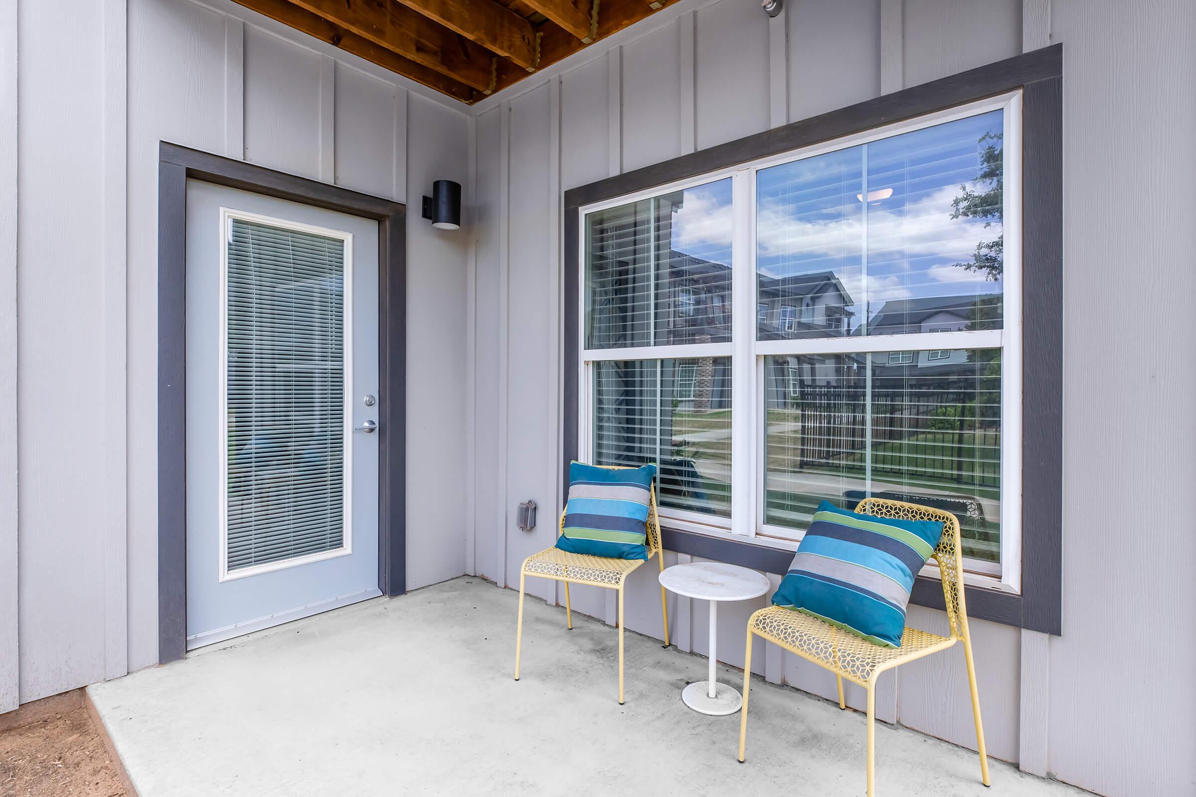 A small outdoor seating area featuring two stylish chairs with blue and green striped cushions, a round white side table, and a door leading inside. The wall is light gray, and there is a window beside the seating, offering glimpses of greenery outside.