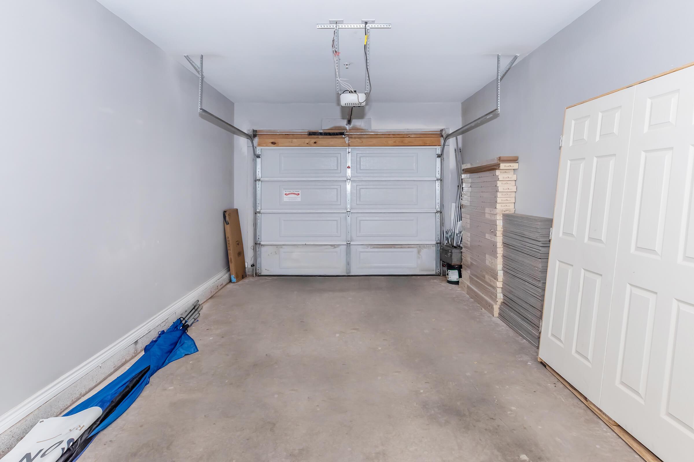 A spacious empty garage featuring a gray concrete floor, a large closed garage door, and a side door. The walls are painted light gray, with some stacked materials on the right side. There's a blue tarpaulin on the floor and minimal items visible, creating a clean and uncluttered appearance.