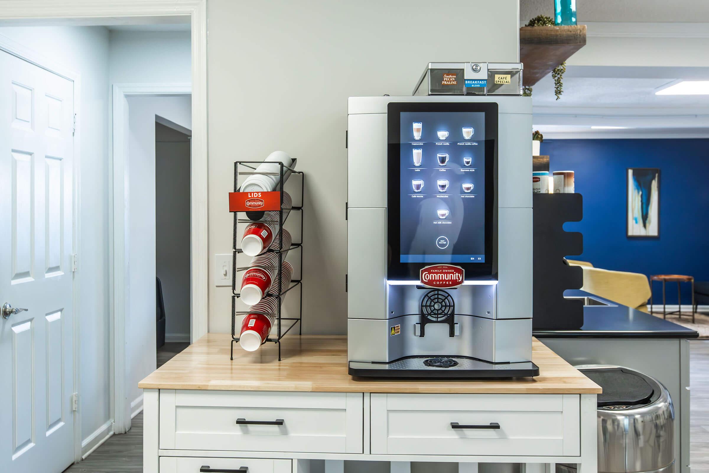 A modern beverage dispenser on a wooden countertop, featuring a digital touchscreen interface. Next to it, a wire rack holds multiple paper cups. The background includes a blue-painted wall with abstract art and light-colored furniture, creating a contemporary office or lounge atmosphere.