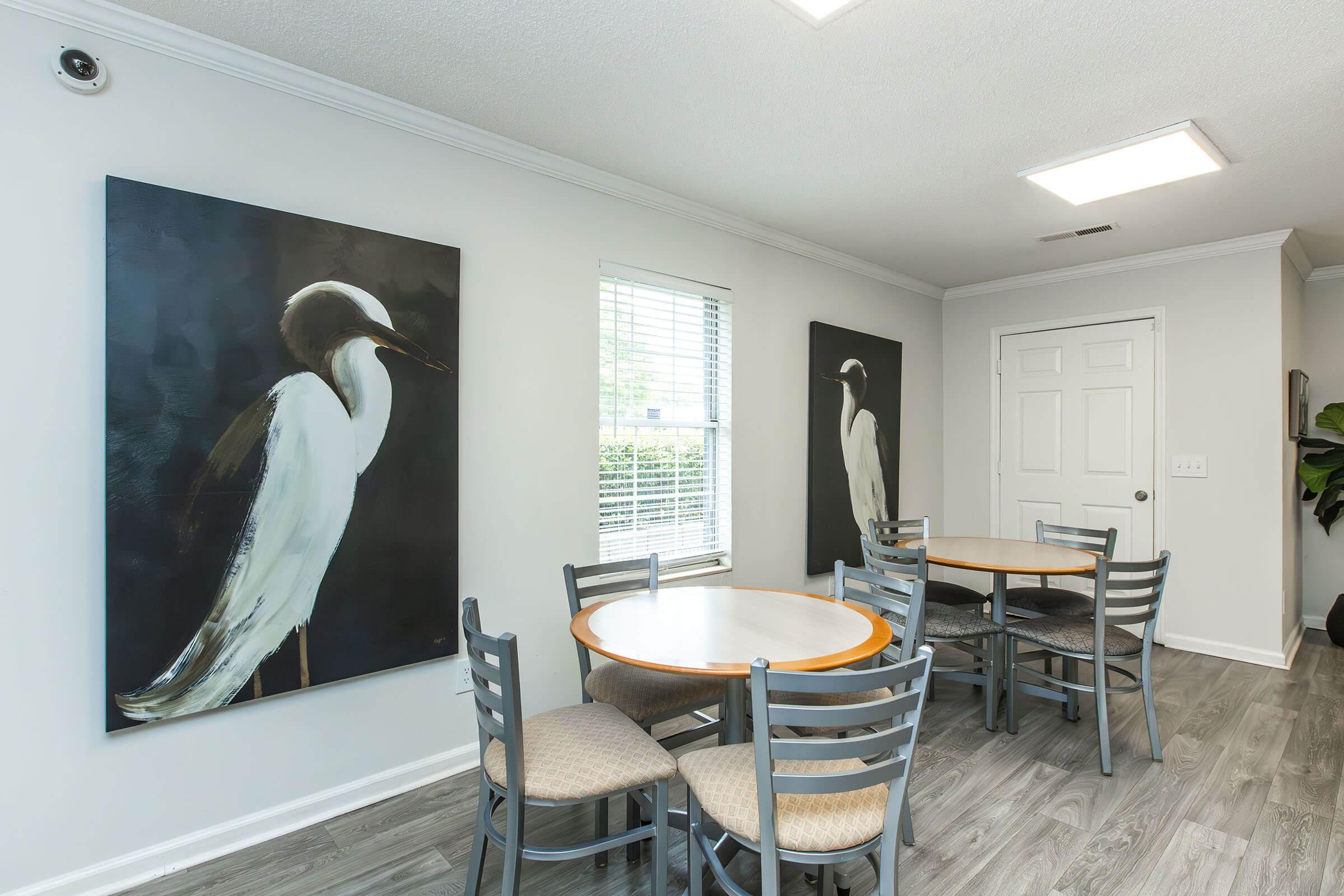 A bright dining area featuring two circular tables surrounded by gray chairs. The walls are adorned with large, artistic paintings of white birds. A window allows natural light to illuminate the space, and there is a door leading to another room. The floor is finished in a light wood-style material.