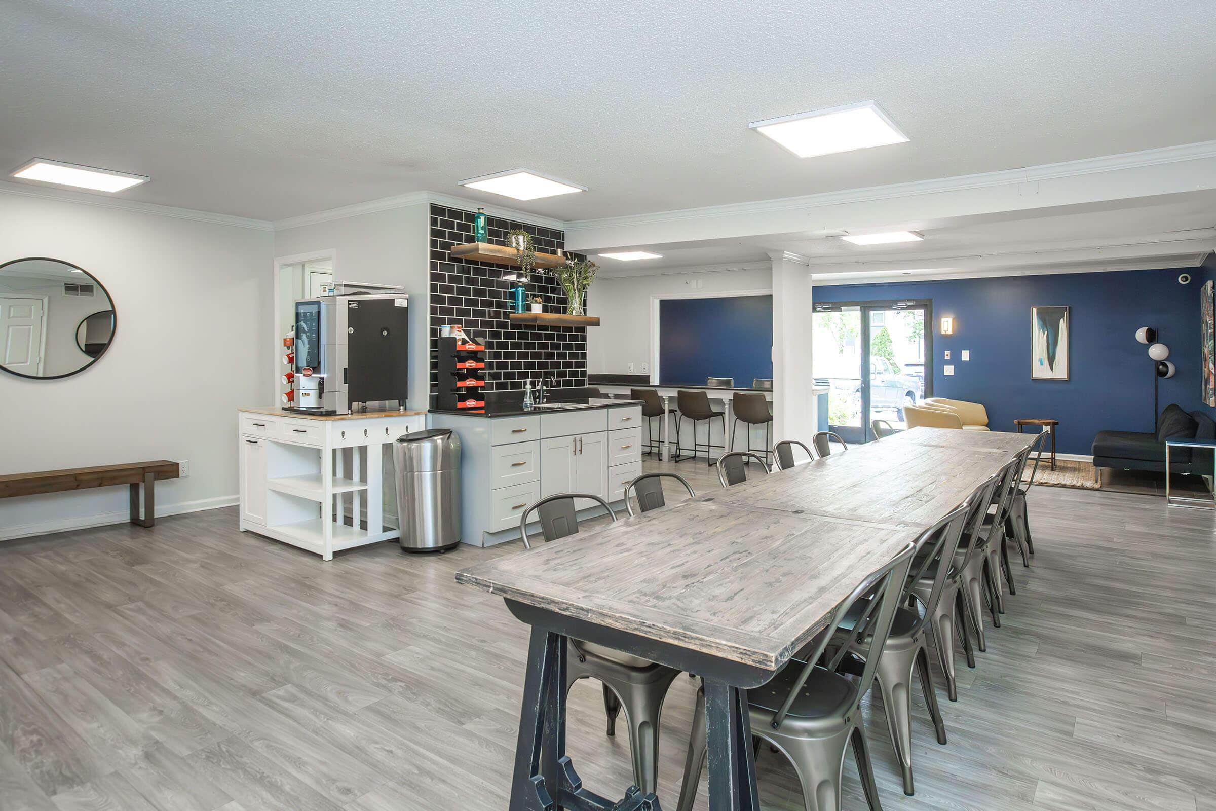 A modern communal kitchen and dining area featuring a long wooden table with metal chairs, a black tile backsplash, and a large window overlooking an outdoor space. The kitchen includes a refrigerator, cabinets, and shelves with decorative items. The walls are painted in neutral and dark tones, creating a stylish atmosphere.