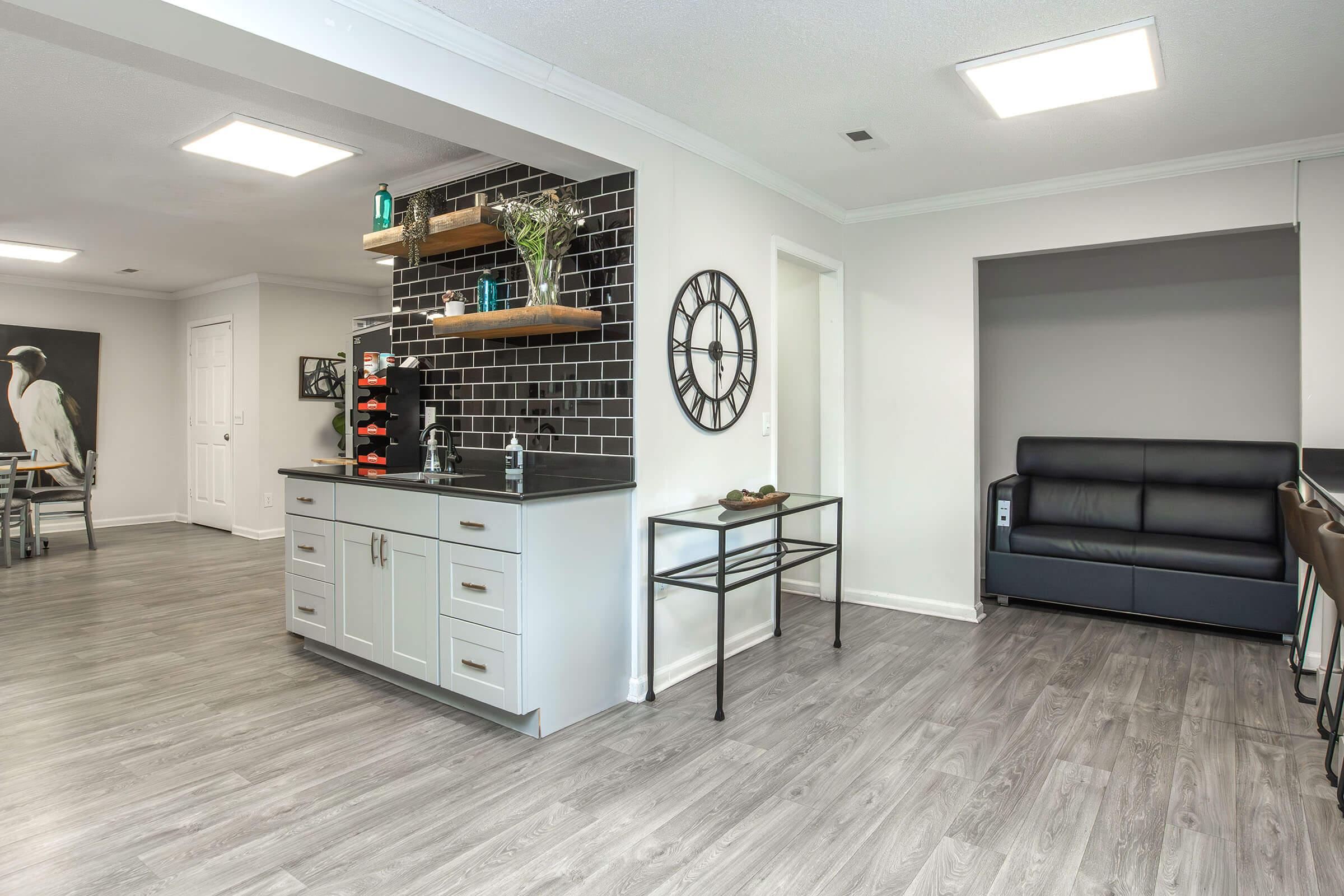 A modern interior view showcasing an open-concept living space. Features include a bar area with shelves holding decorative items, a dining area with a table and chairs, a clock on the wall, and a sleek black couch in the adjoining room. The flooring is light-colored with a contemporary vibe.
