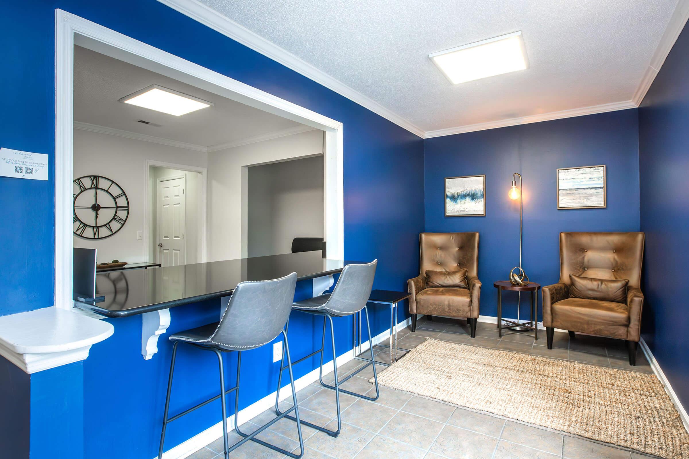A modern interior featuring a seating area with two brown leather chairs and a bar counter with three stylish stools. The walls are painted a bold blue, complemented by a round wall clock and two framed artworks. A soft area rug lies on the tiled floor, enhancing the contemporary aesthetic of the space.