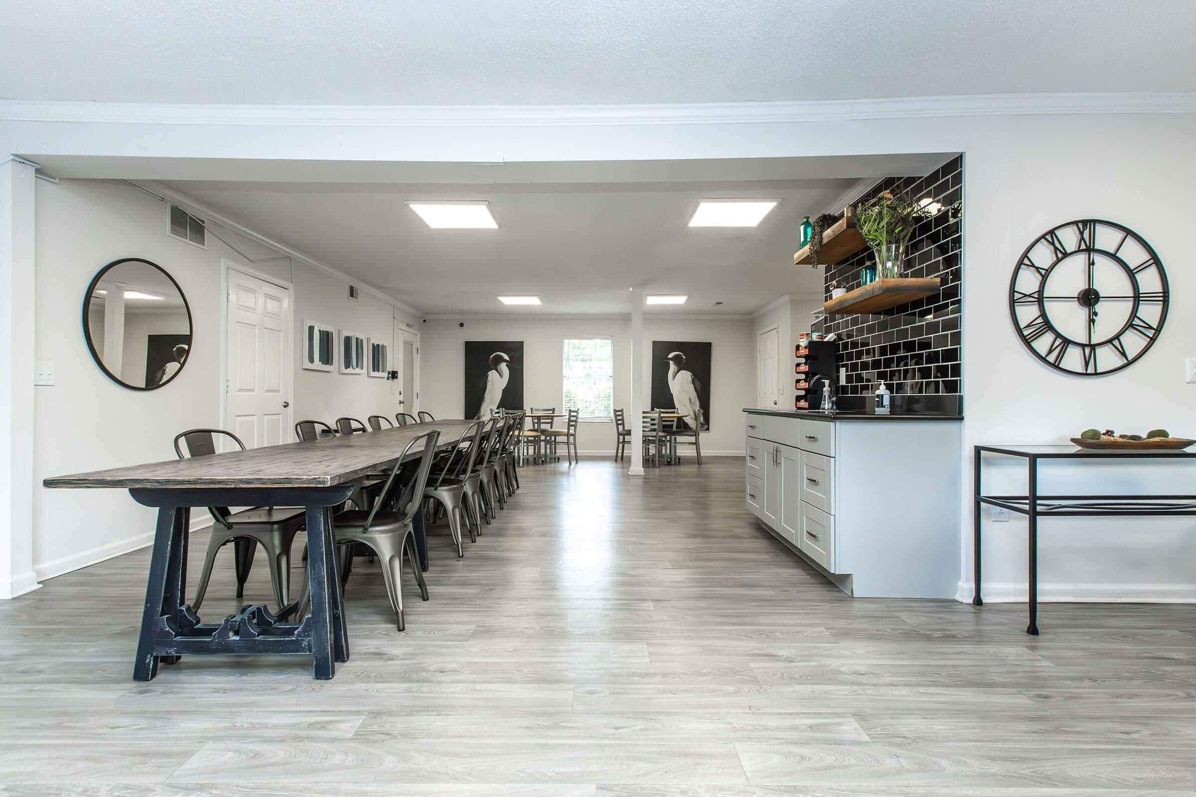 A spacious, modern dining area featuring a long wooden table with metal chairs, gray walls, and large mirrors. On one side, there's a black and white patterned wall with clocks and decorative items. Large windows provide natural light, and minimalistic decorations create a clean, inviting atmosphere.