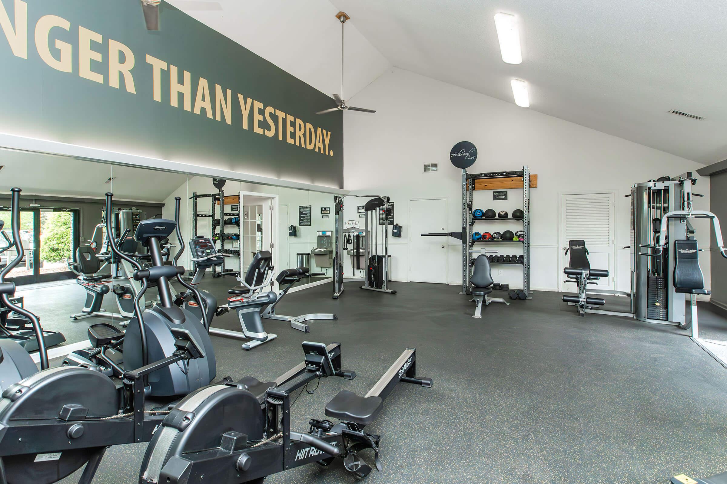 A gym interior featuring various exercise machines, including rowing machines and weight benches. The space has a bright, open feel with large windows, a ceiling fan, and motivational wall art that reads "Stronger than Yesterday." Fitness equipment is neatly arranged along the walls.