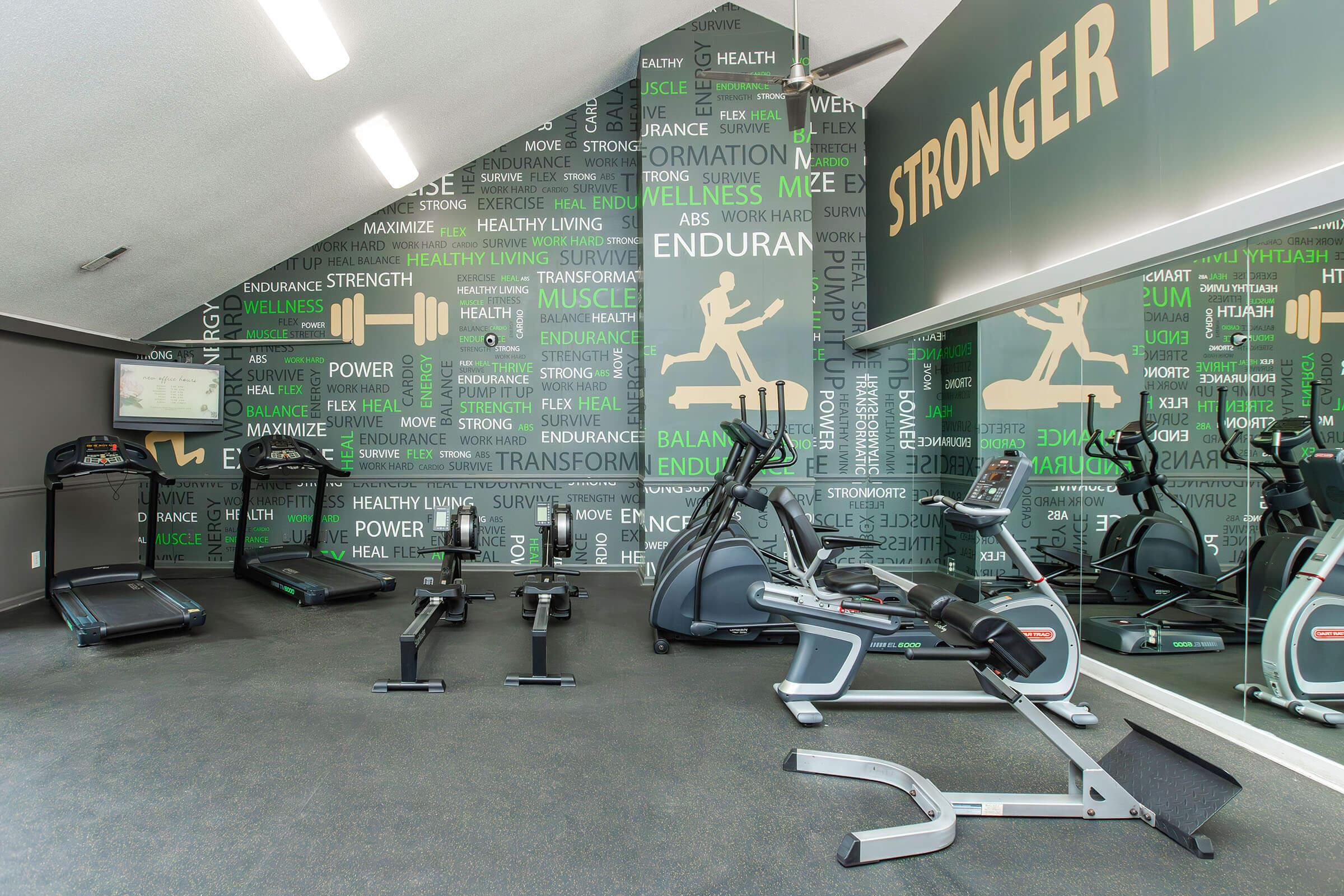 Fitness center interior featuring exercise equipment including treadmills, a rowing machine, and an elliptical. The walls are decorated with motivational phrases related to health, strength, and wellness, creating an energizing atmosphere for workouts. Mirrors enhance the spacious feel of the gym.
