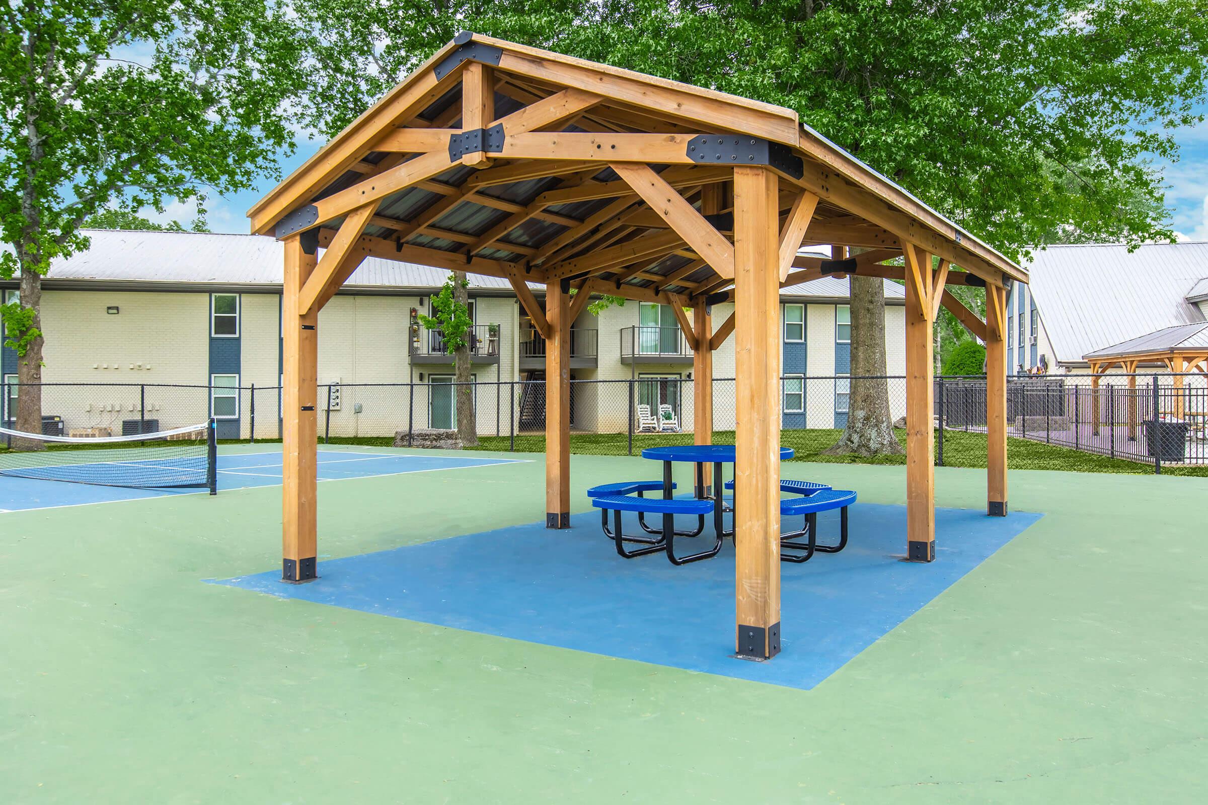 A wooden gazebo with a pitched roof and blue picnic tables underneath, situated on a green and blue court area. In the background, there are buildings with windows and a fenced tennis court. The scene is bright with a clear sky and lush trees nearby.