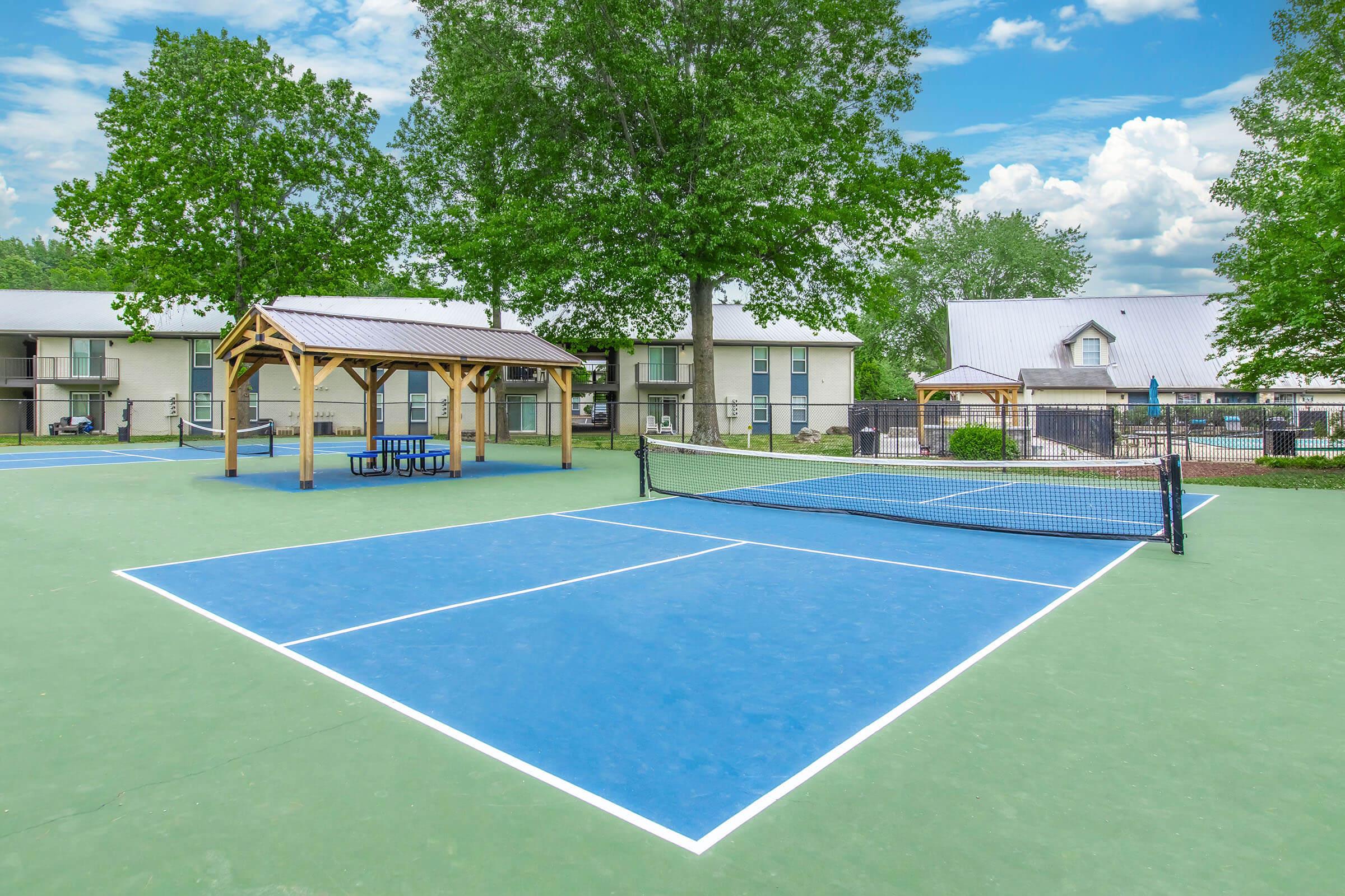 Tennis court with vibrant blue and green surfaces, surrounded by trees and residential buildings. A covered gazebo with seating is nearby, and the sky features scattered clouds. The setting is bright and inviting, suggesting an outdoor recreational area.