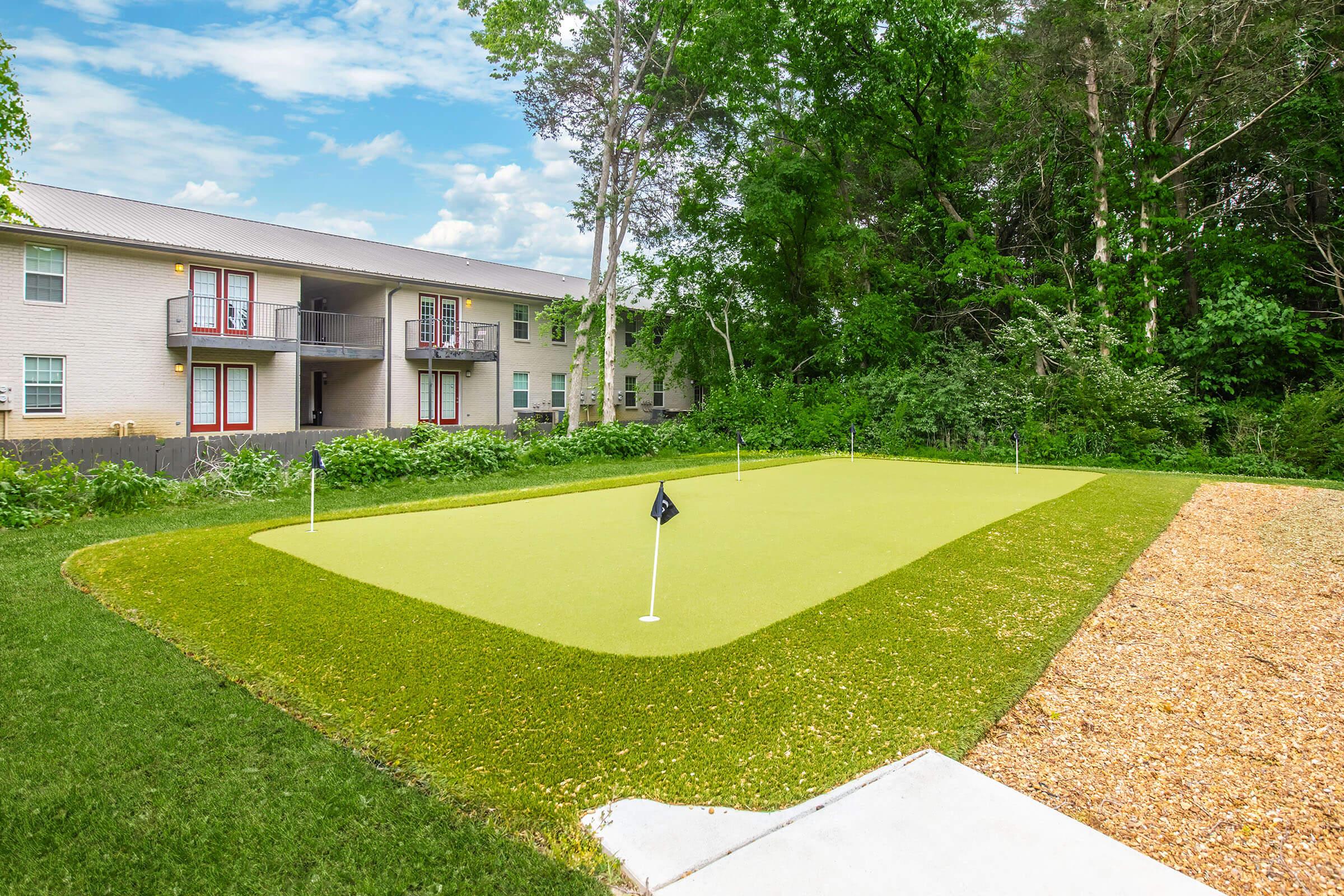 A well-maintained putting green with two flags, surrounded by lush greenery and trees. In the background, a multi-story apartment building with balconies is visible. The area features a pathway leading to the green and gravel on one side, creating a serene recreational space.