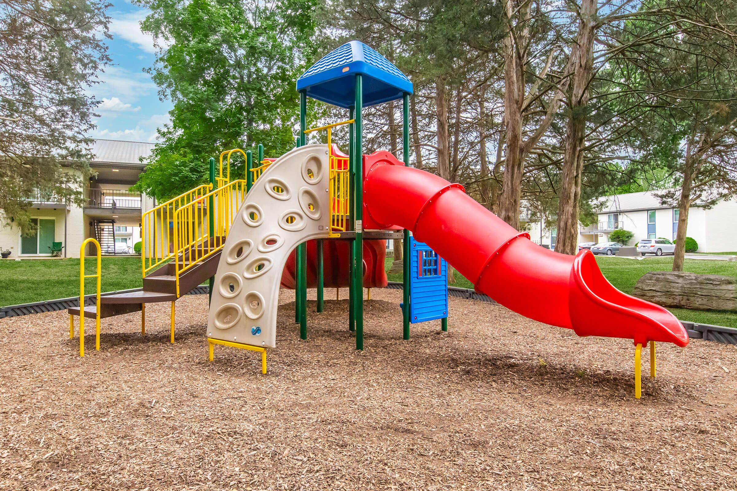 A colorful playground structure featuring a yellow climbing wall, a blue rooftop, and a large red slide. The playground is surrounded by soft wood chip flooring and greenery in the background. Perfect for children to play and explore.