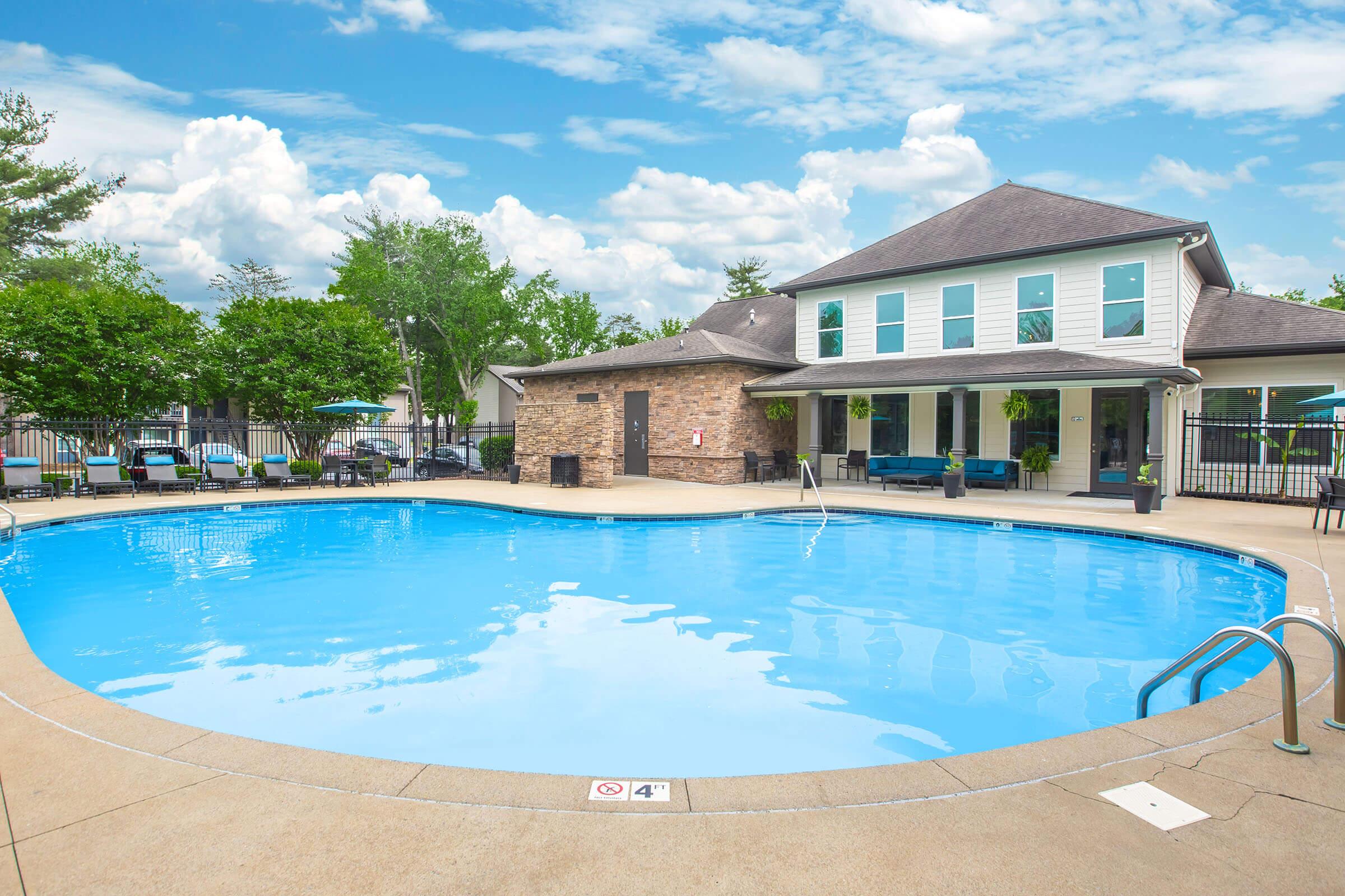 A clear blue swimming pool surrounded by a concrete deck. In the background, a modern building with large windows and a stone exterior. Lush green trees and blue skies with scattered clouds enhance the serene atmosphere. Poolside furniture is visible, creating a welcoming leisure space.