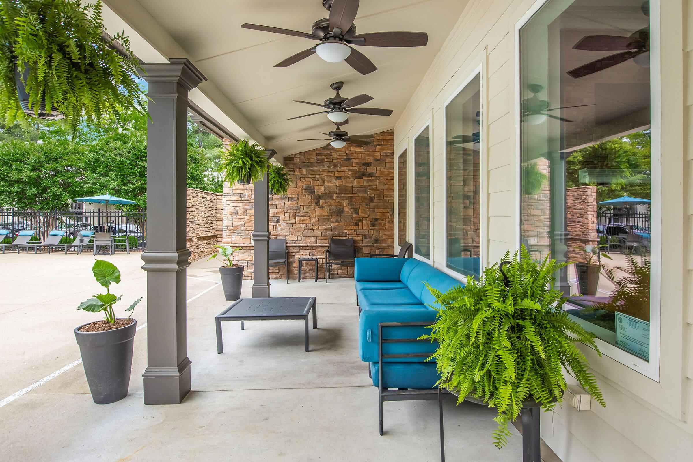 A covered patio area featuring a blue sofa and coffee table, flanked by decorative ferns in planters. Stone and wood accents are visible on the walls, with ceiling fans overhead. The background shows a pool area with lounge chairs.