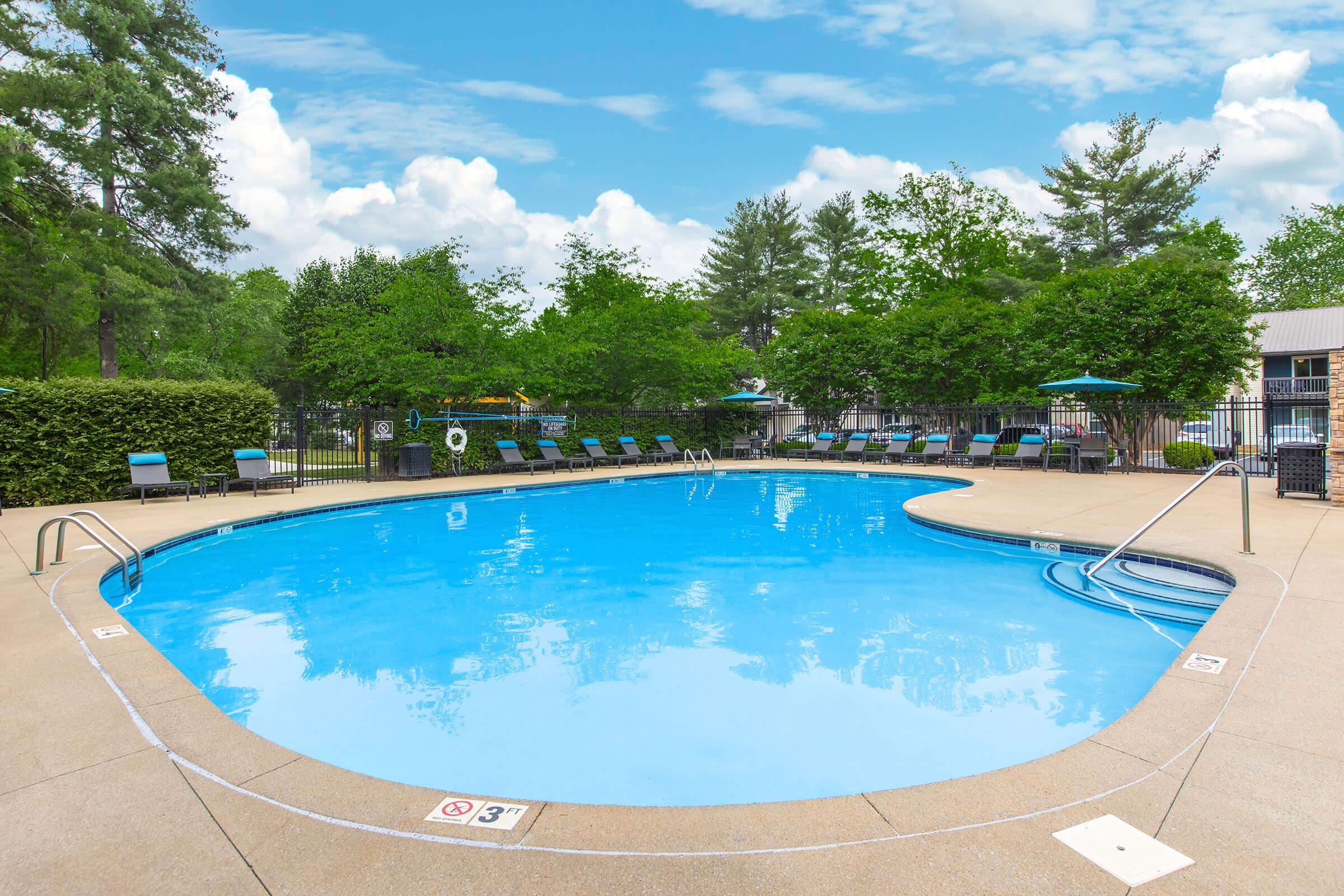 A clean, inviting outdoor swimming pool surrounded by green trees and lounge chairs. The pool features clear blue water, a gentle slope in one area, and is bordered by a fence. In the background, there are umbrellas for shade, and the sky is partly cloudy with a bright blue tone.