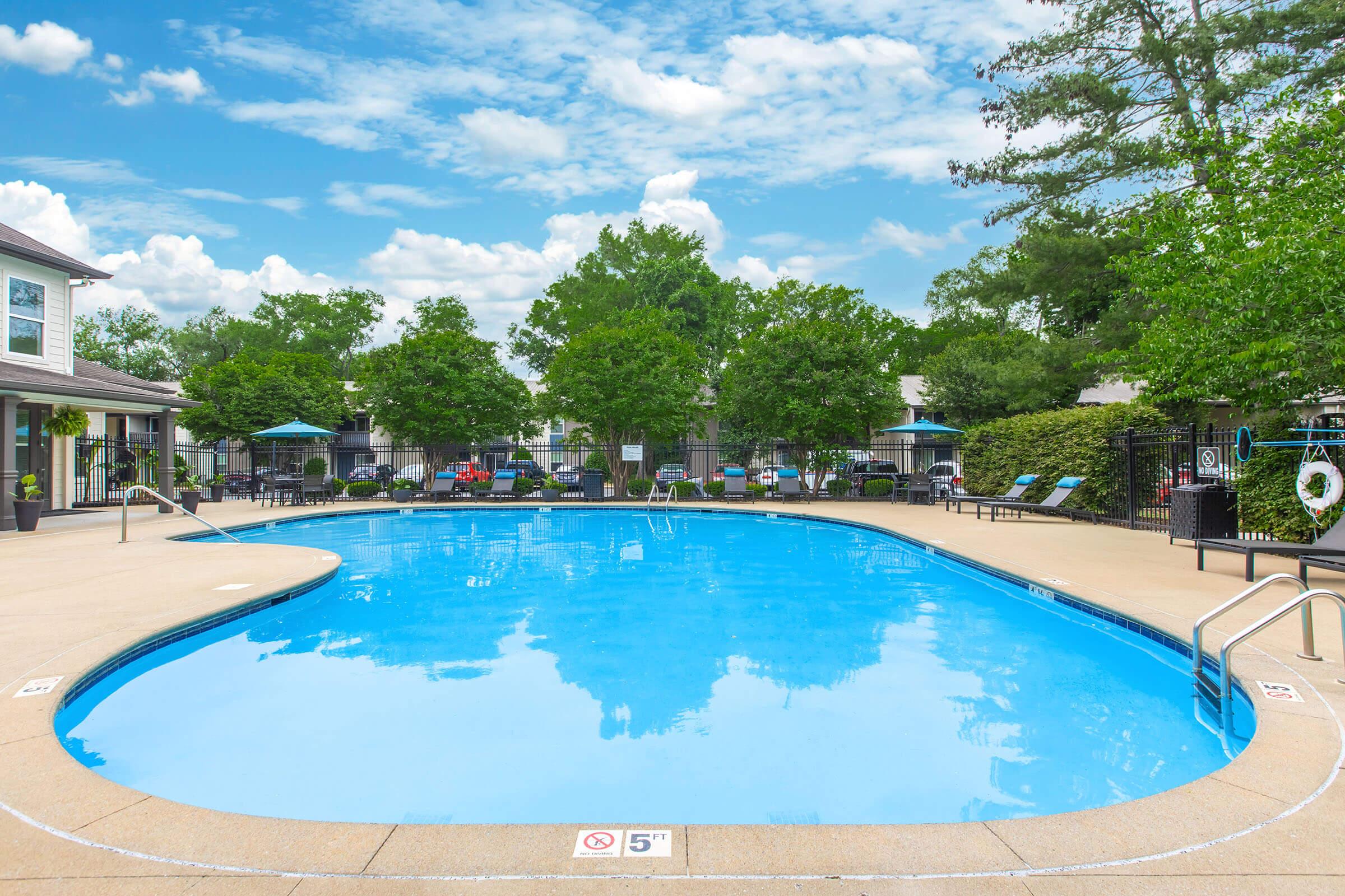 A bright outdoor swimming pool with crystal-clear water, surrounded by lush green trees and lounge chairs. The sky is partly cloudy, creating a serene atmosphere. There's a shallow end marked with a depth of 5 feet, and several umbrellas are placed near the pool area for shade.