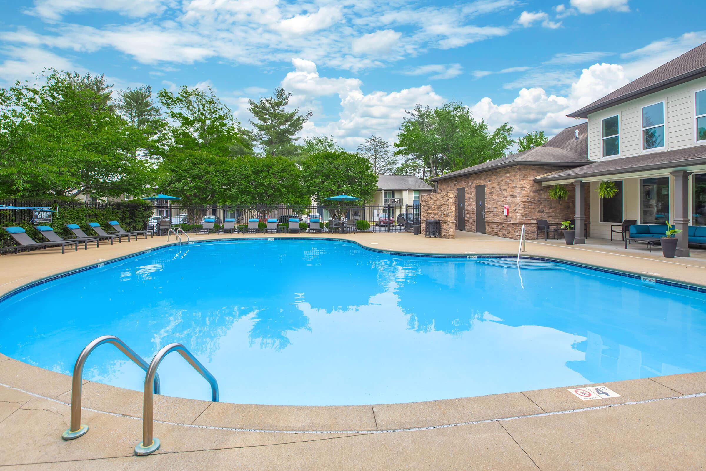 A clear blue swimming pool surrounded by lounge chairs and green trees. The pool area features a stone building with seating and umbrellas, under a bright, partly cloudy sky. The scene conveys a relaxing outdoor atmosphere, perfect for leisure and enjoyment.