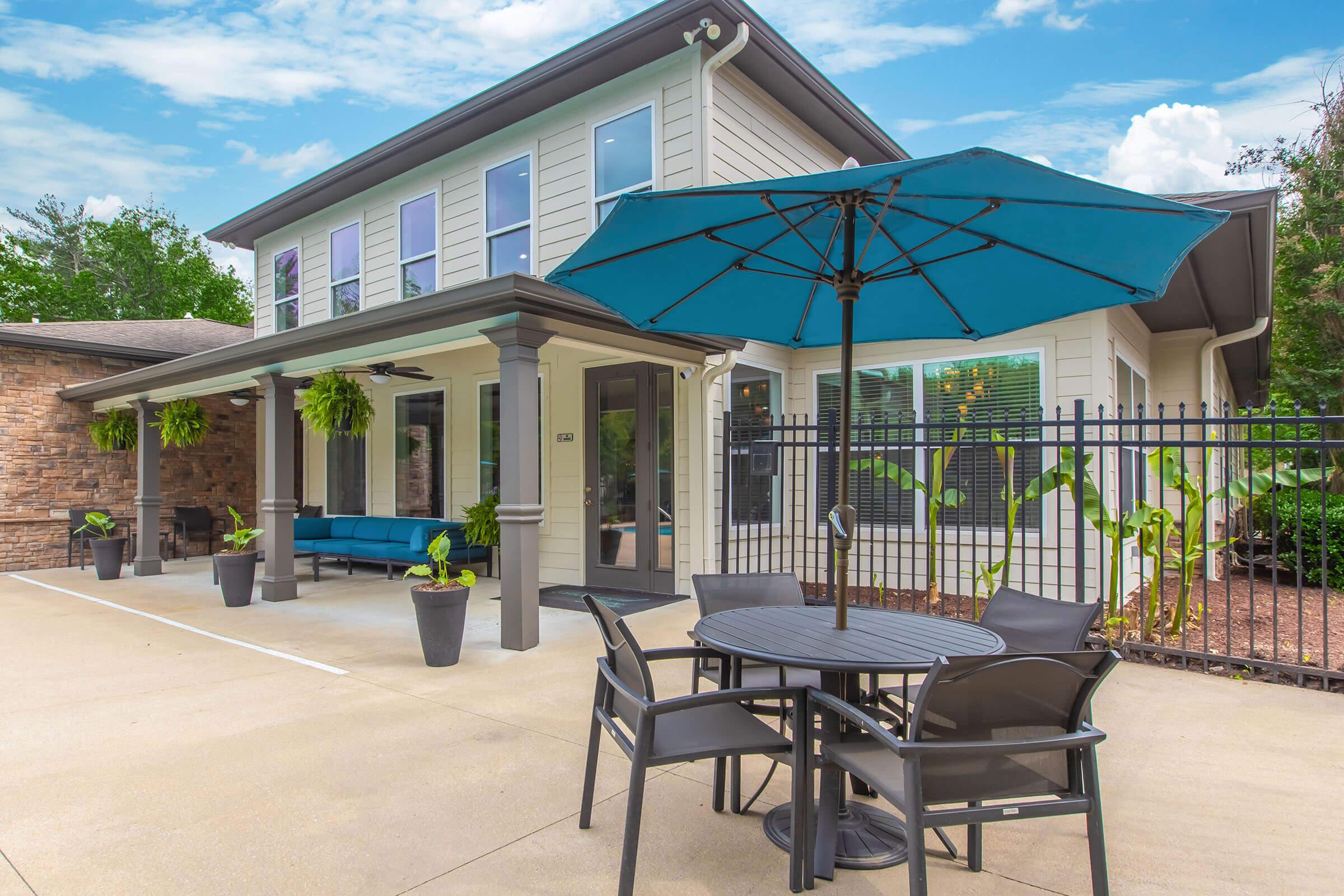 A modern building exterior with a patio area featuring a black table and chairs under a blue umbrella. The space includes potted plants and a comfortable seating area with a blue sofa. The design is clean and inviting, set against a background of greenery and a clear blue sky.