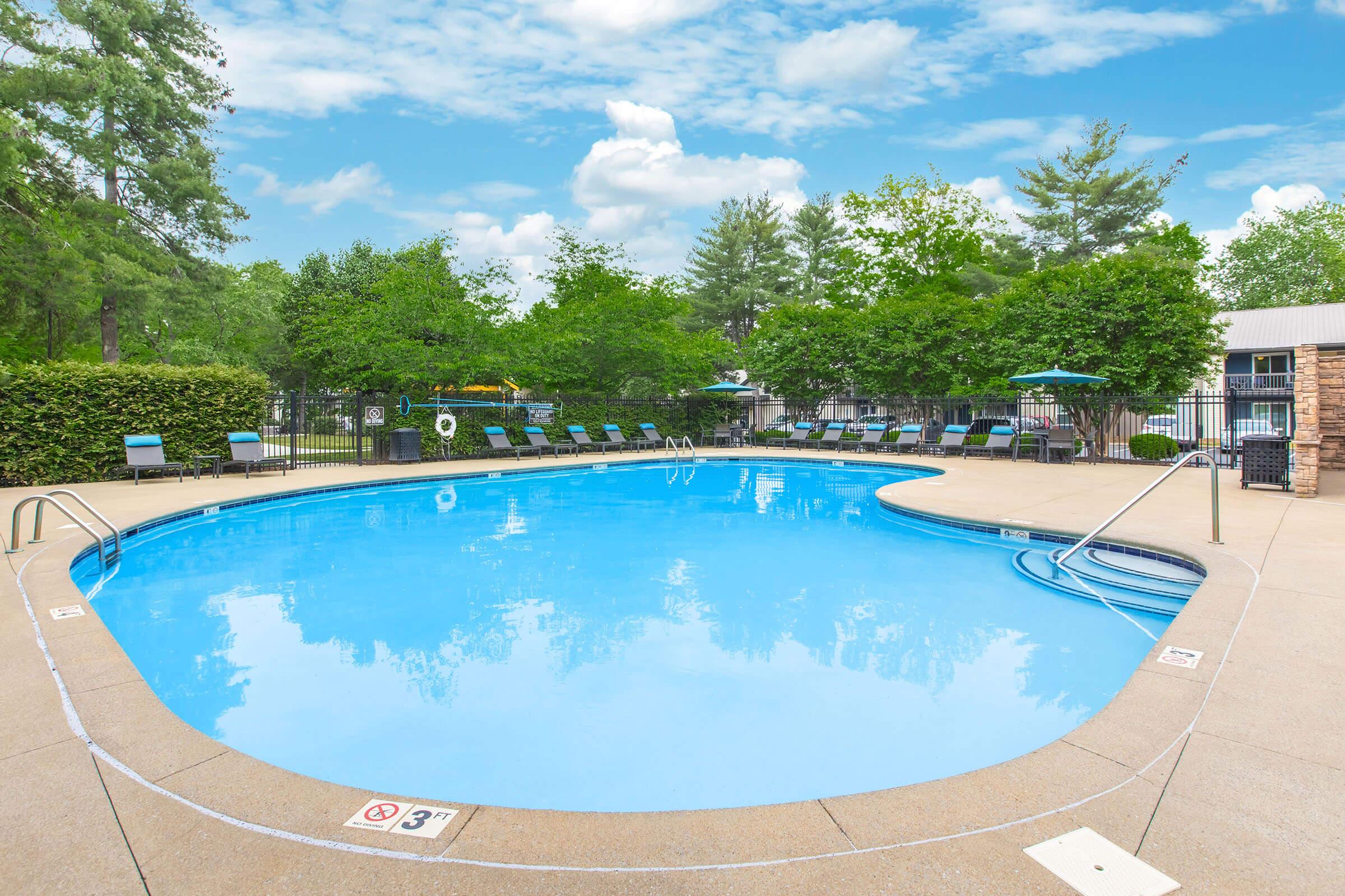 Clear blue swimming pool surrounded by green trees and neatly arranged lounge chairs. The pool features a shallow end marker and a deck area with umbrellas for shade. A bright sky with scattered clouds completes the scenic outdoor setting.