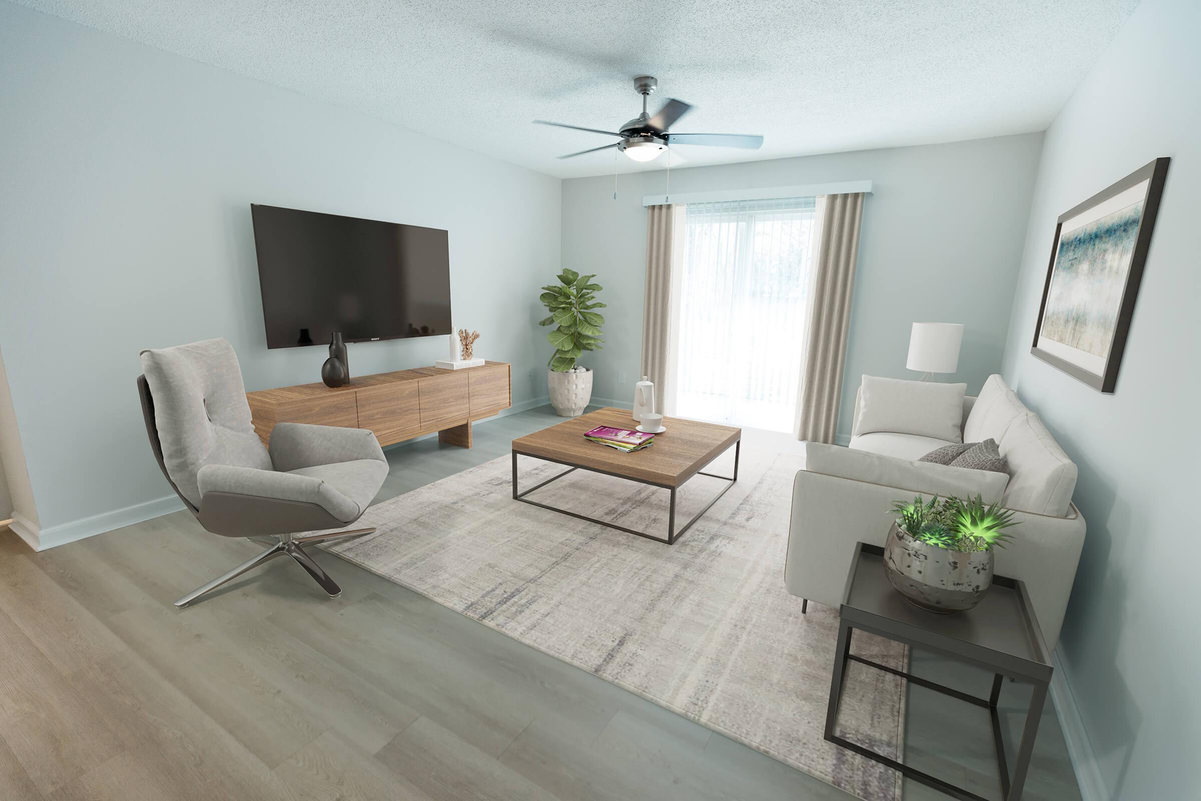 A modern living room featuring a light blue wall, a large flat-screen TV on a wooden media console, a comfortable gray armchair, and a cozy white sofa. A coffee table is in the center with magazines on it. A potted plant adds greenery, and large windows with sheer curtains let in natural light.
