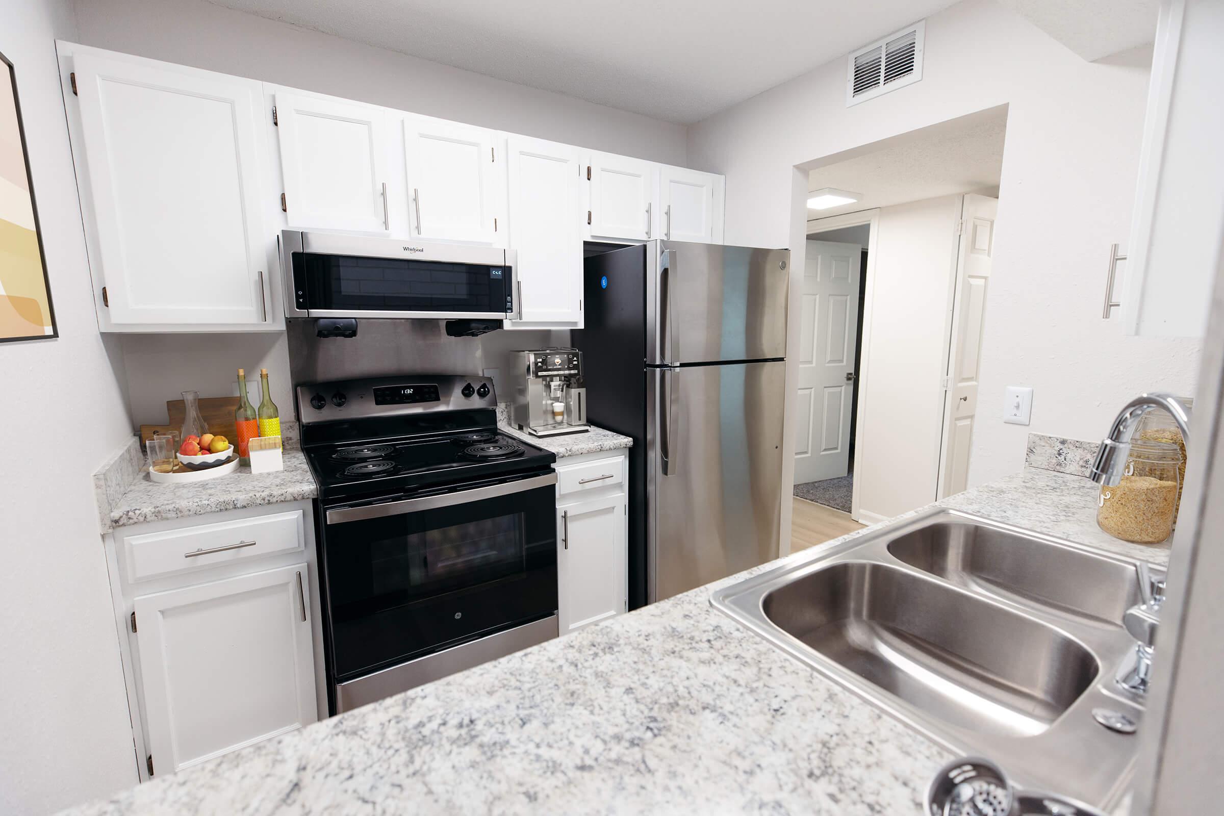 A modern kitchen featuring white cabinets, a stainless steel refrigerator, and an electric stove. The countertop is made of light-colored granite. There’s a bowl of fruit on the counter, and a sink with two basins is visible. The walls are painted light colors, creating a bright and inviting atmosphere.