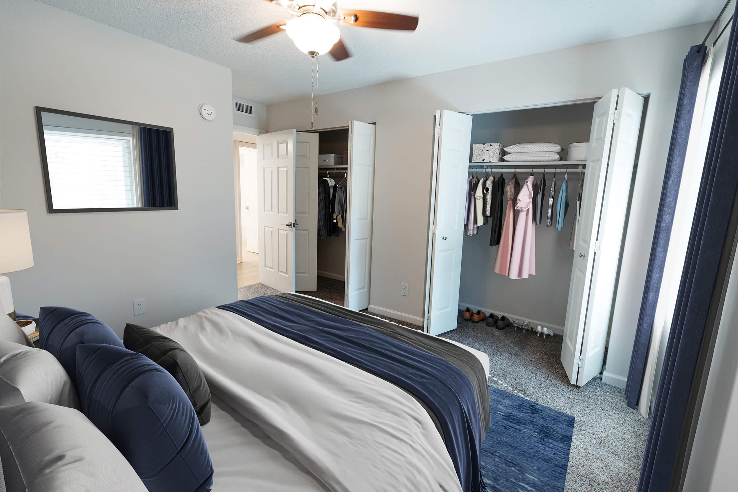A modern bedroom featuring a neatly made bed with blue and white bedding, a bedside lamp, and a ceiling fan. Two open closet doors reveal organized clothing and accessories. Light-colored walls and gray carpet enhance the airy feel of the space. A doorway in the background leads to another room.