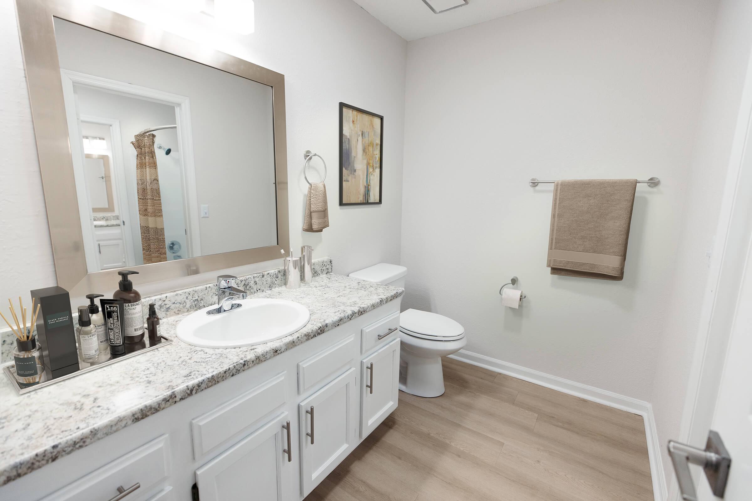 A modern bathroom featuring a white sink with a granite countertop, a mirror framed in gold, beige towels hanging on a towel rack, and a decorative wall art piece. The space includes a toilet and neutral-colored flooring, creating a clean and inviting atmosphere.
