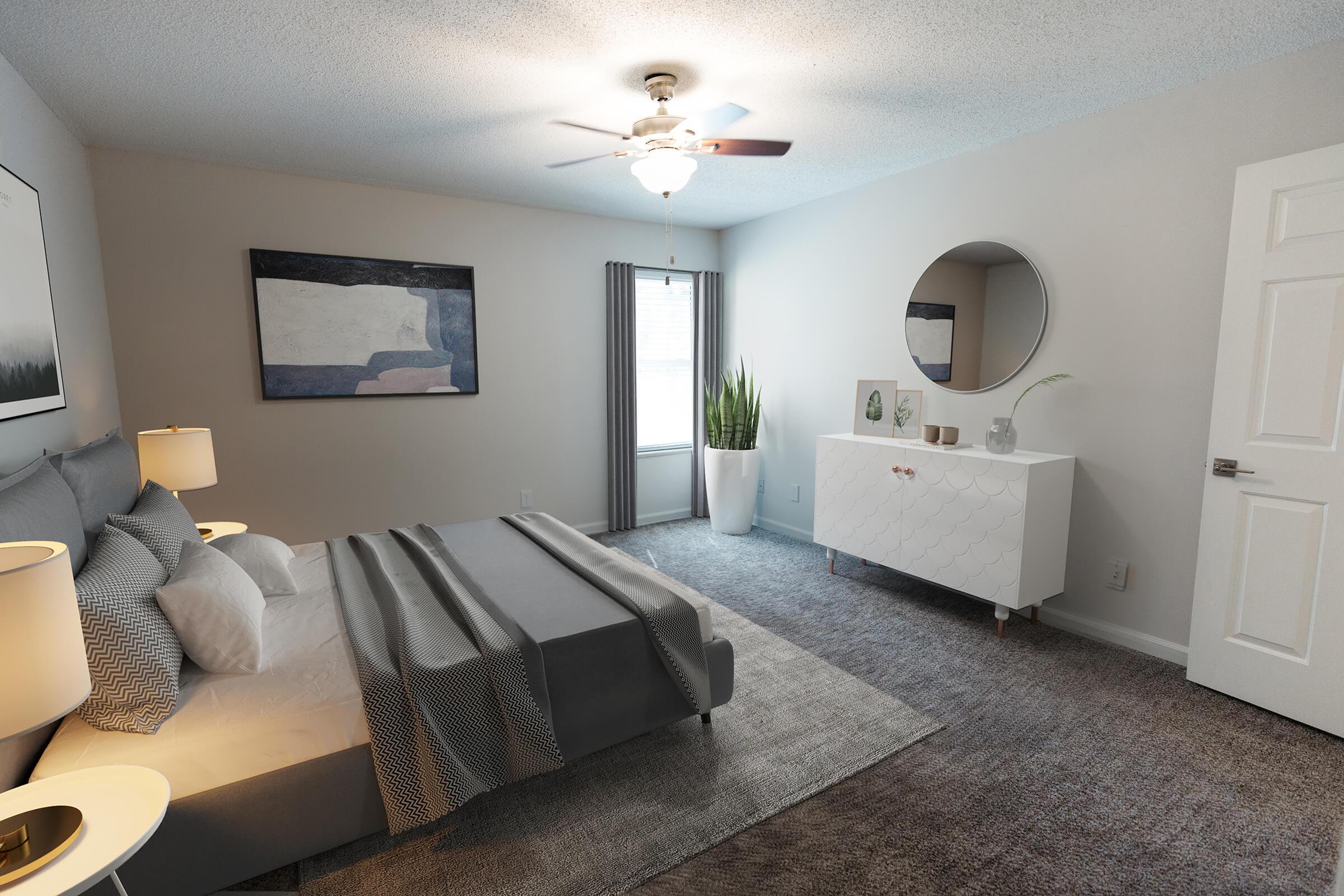 A modern bedroom featuring a queen-sized bed with gray bedding, nightstands with lamps, and a decorative area rug. There is a white dresser with circular mirror, a tall indoor plant by the window, and artwork on the wall. The room has neutral tones and a ceiling fan, creating a cozy atmosphere.