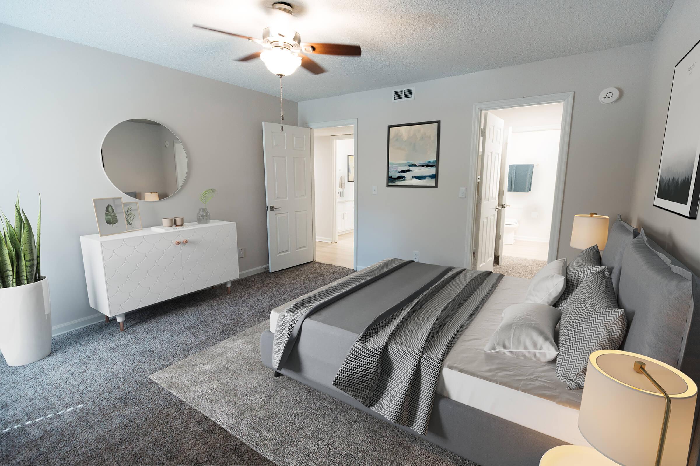 A bright, modern bedroom featuring a bed with gray bedding, a sleek nightstand with a lamp, and a decorative mirror on the wall. A potted plant stands in the corner, and two doors lead to adjacent rooms, suggesting a spacious and inviting atmosphere.