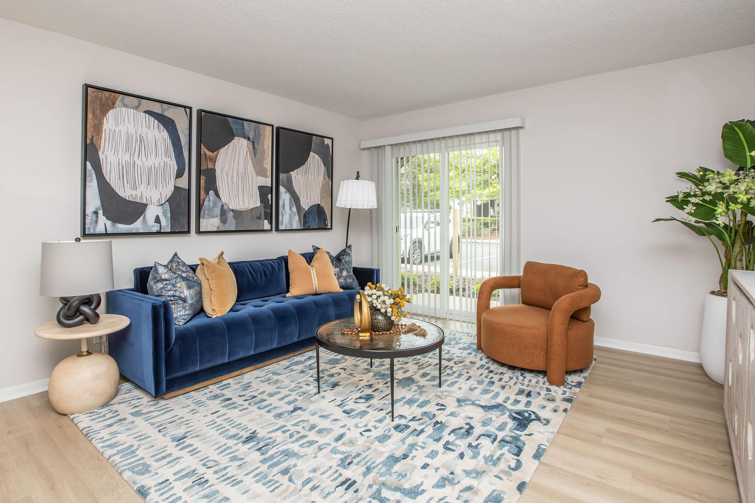A modern living room featuring a blue velvet sofa adorned with decorative pillows, a round glass coffee table, and an orange accent chair. Artwork with abstract designs hangs on the wall. A lamp next to the sofa provides soft lighting, and a large window allows natural light to illuminate the space, complemented by greenery.