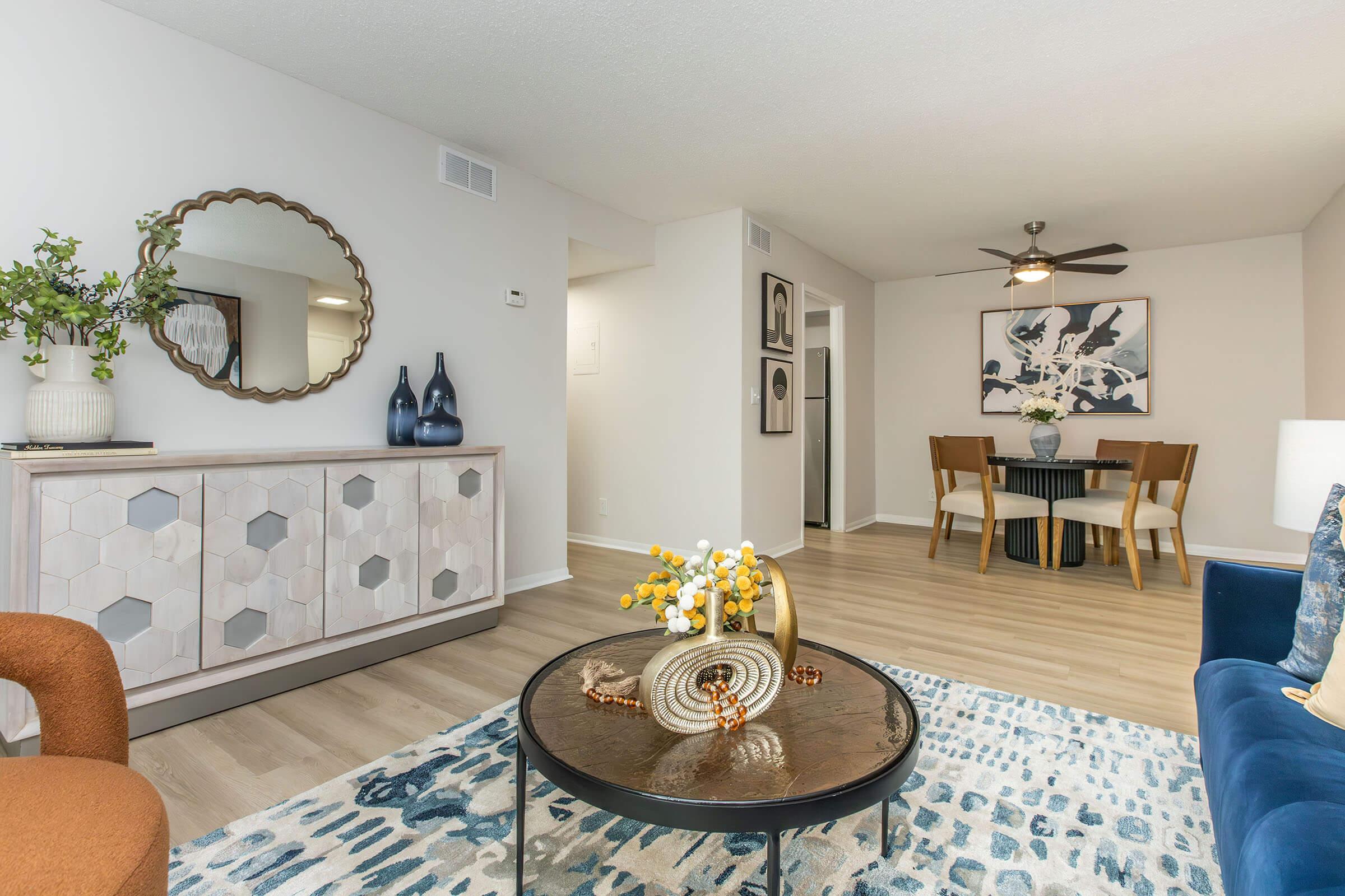 A modern living room featuring a circular mirror above a stylish dresser, decorative vases, a coffee table with a floral arrangement, and a blue sofa. Light wooden flooring complements the neutral walls, and there is a dining area with a round table and chairs in the background, illuminated by a ceiling fan.