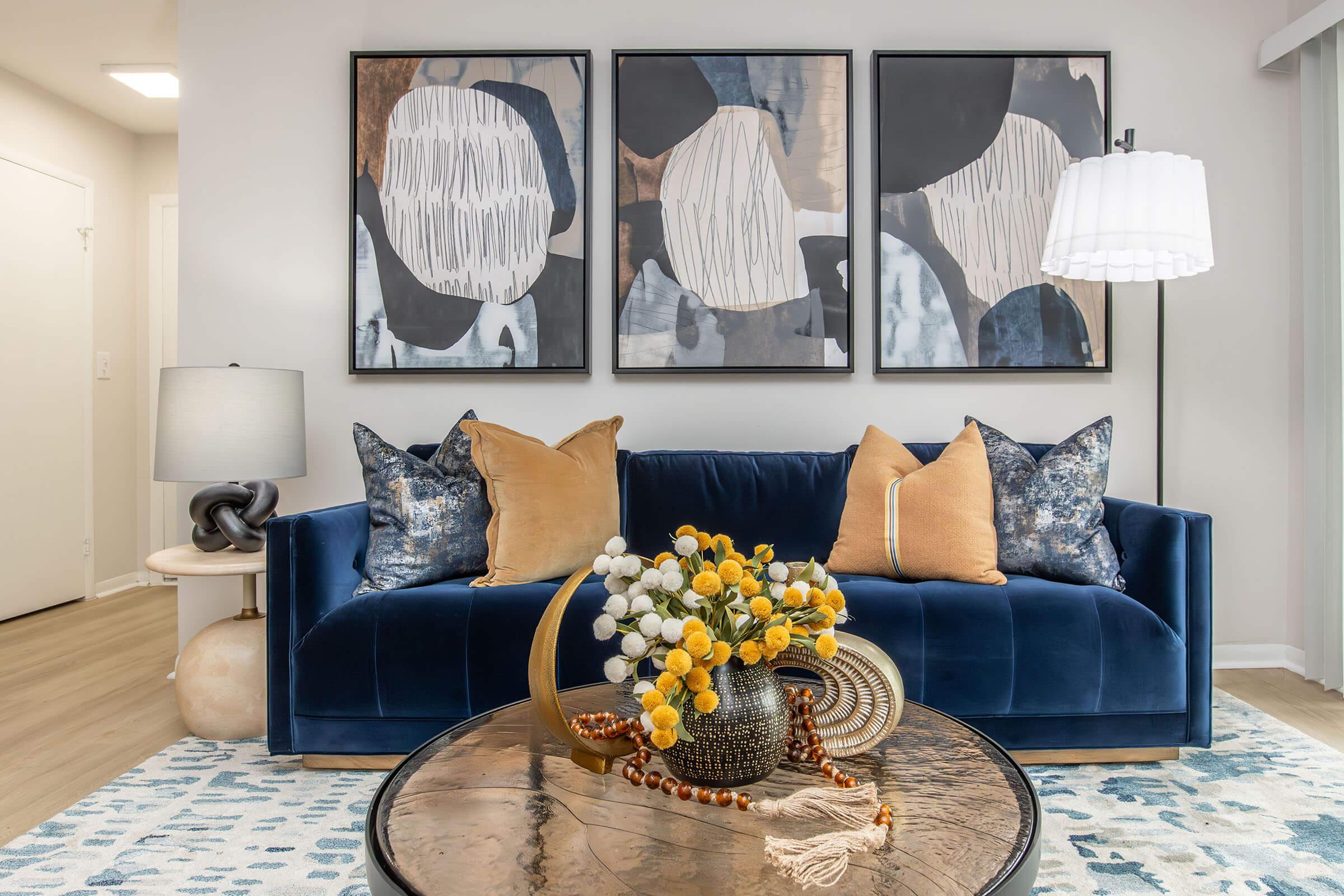 A modern living room featuring a navy blue sofa adorned with patterned throw pillows in shades of orange and blue. A round coffee table with a decorative arrangement of flowers and beads sits in front. Three abstract framed artworks hang on the wall, and a stylish floor lamp stands beside the sofa.
