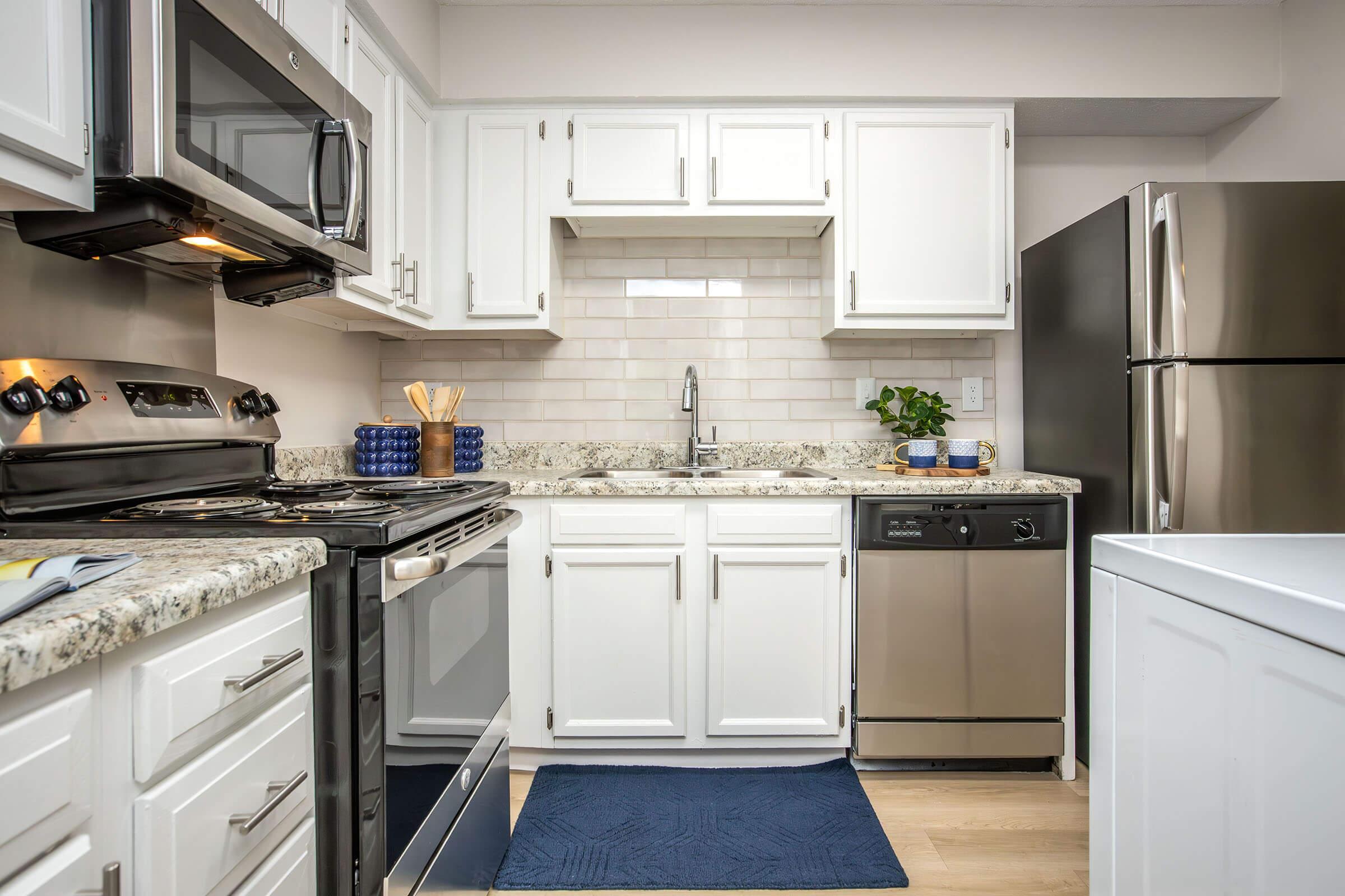 Modern kitchen featuring white cabinetry, stainless steel appliances including a microwave, oven, refrigerator, and dishwasher. The countertop is made of speckled granite, and there's a sink with a faucet. A blue area rug is on the floor, and kitchen utensils are displayed in a holder on the countertop.