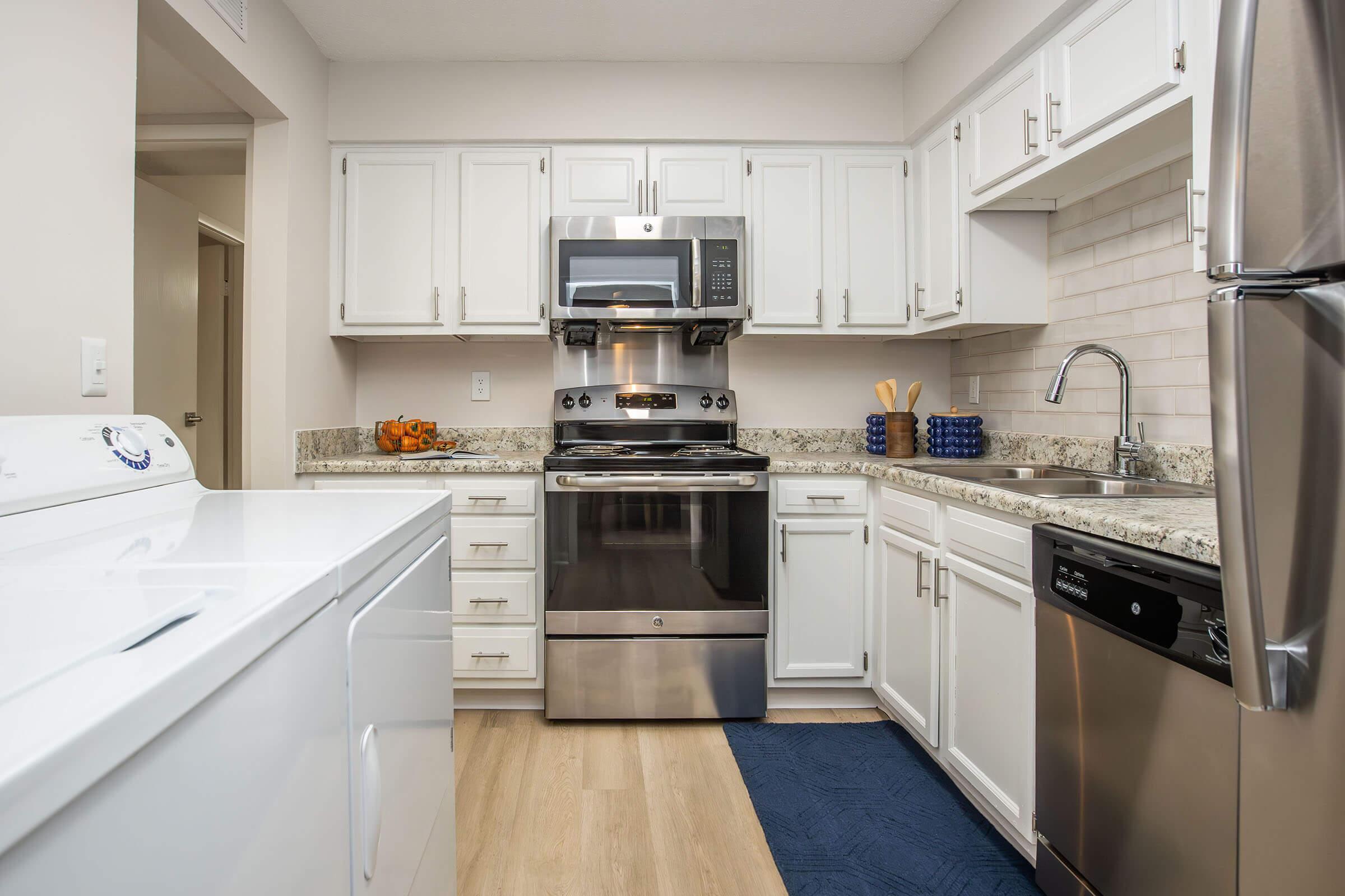A modern kitchen featuring white cabinets, a stainless steel oven and microwave, a sink with a faucet, and a dishwasher. There is a laundry machine on the left and a countertop with granite finish. A bowl of oranges is visible on the counter, and the floor is light wood with a blue mat.