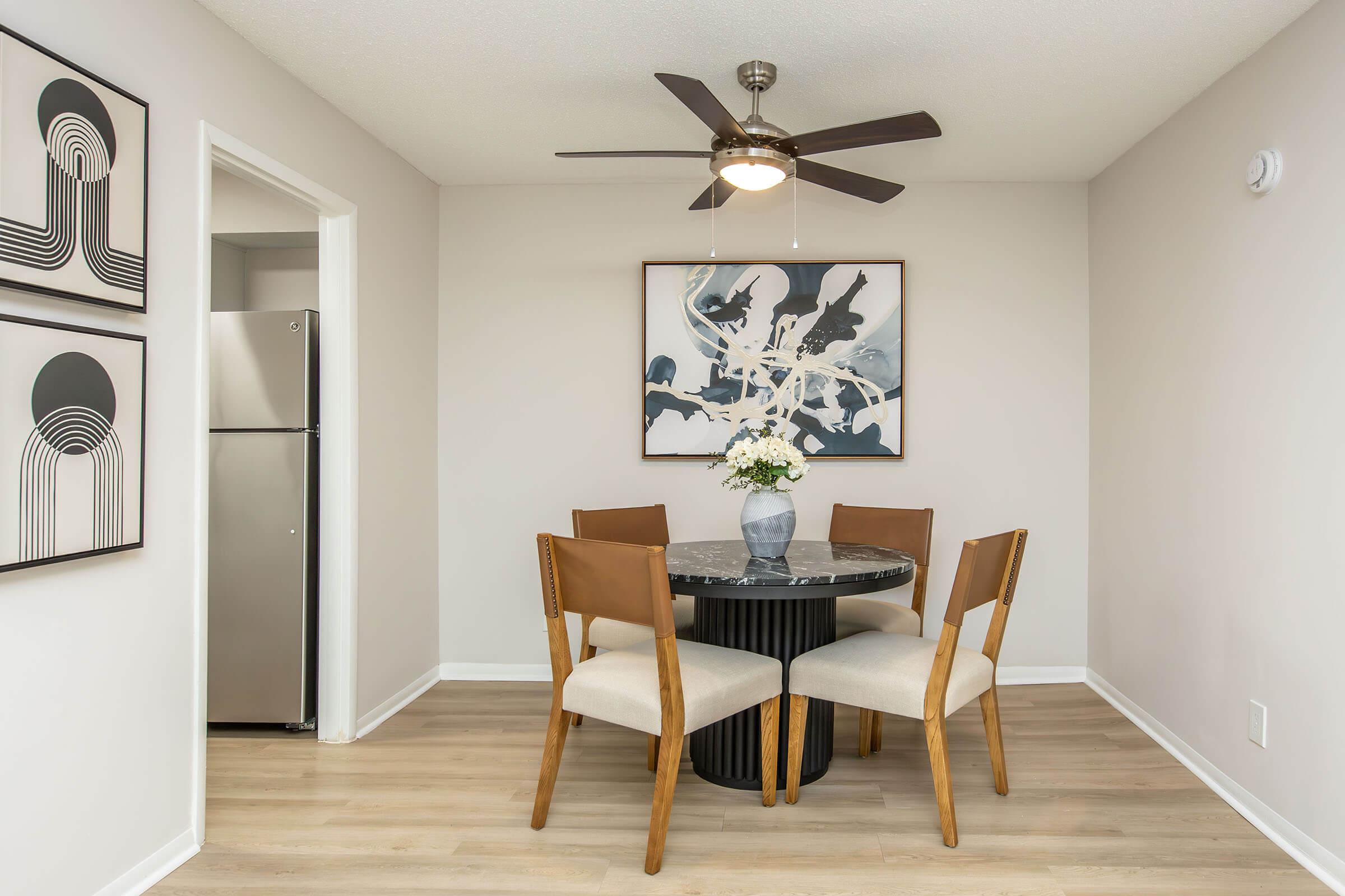 A cozy dining area featuring a round black table with four wooden chairs. A vase of flowers sits on the table. The walls are decorated with two abstract art pieces, and there's a door leading to a small kitchen visible in the background, complete with a stainless steel refrigerator. A ceiling fan is also present.