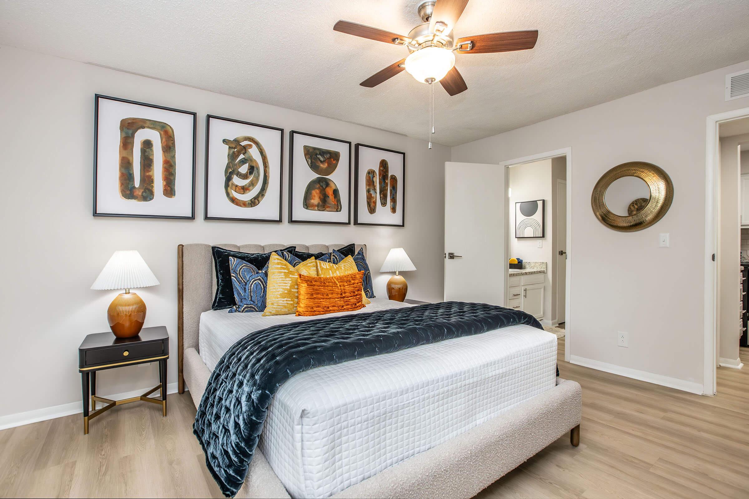 A modern bedroom featuring a neatly made bed with decorative pillows, two lamps on bedside tables, and framed artwork on the wall. The room has light wood flooring, a ceiling fan, and an open door leading to a bathroom area, creating a warm and inviting atmosphere.