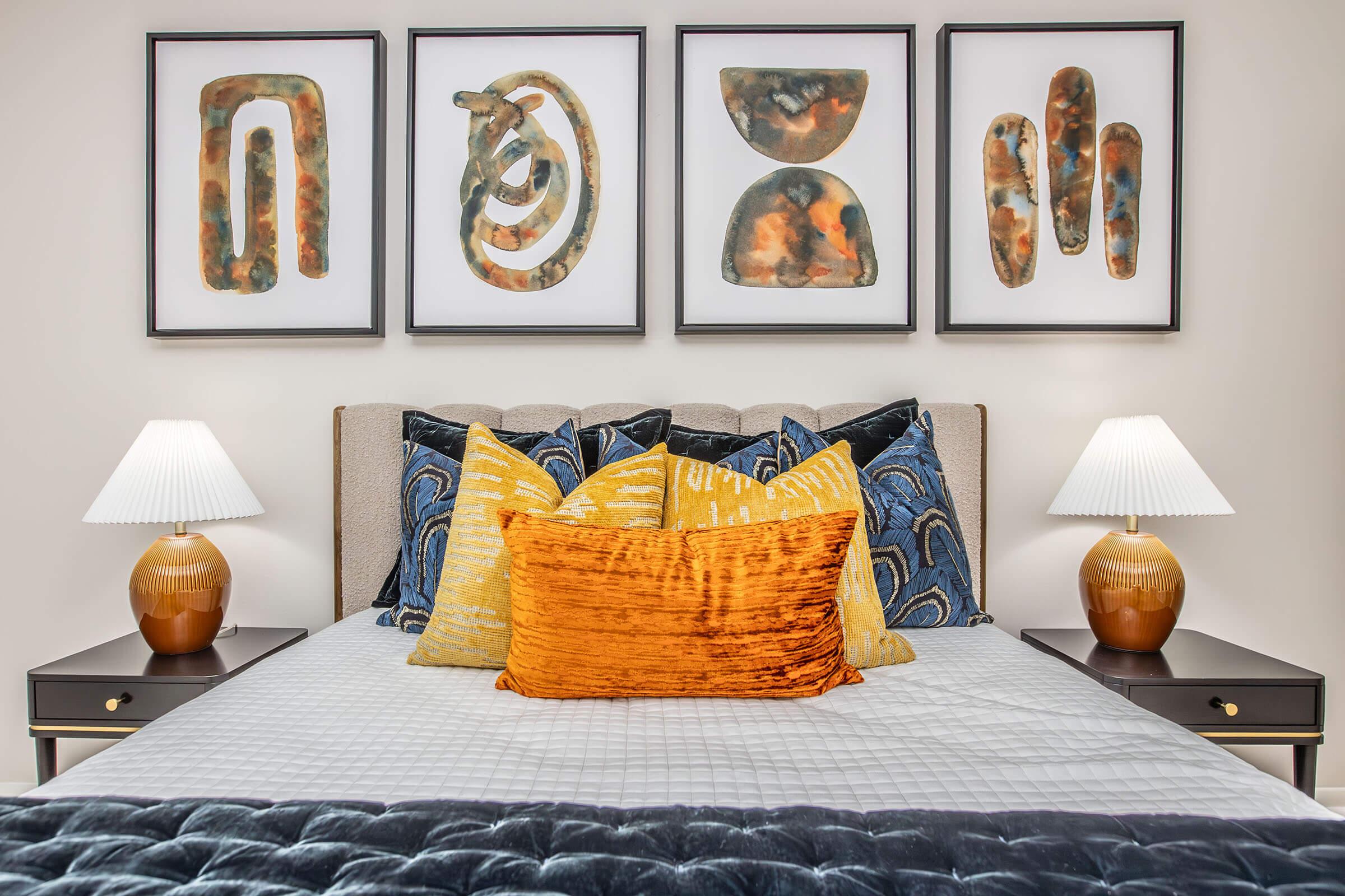 A stylish bedroom featuring a neatly made bed with decorative pillows in various colors and textures. Above the bed are four framed abstract art pieces. On either side of the bed, there are modern nightstands with elegant lamps, creating a cozy and inviting atmosphere.