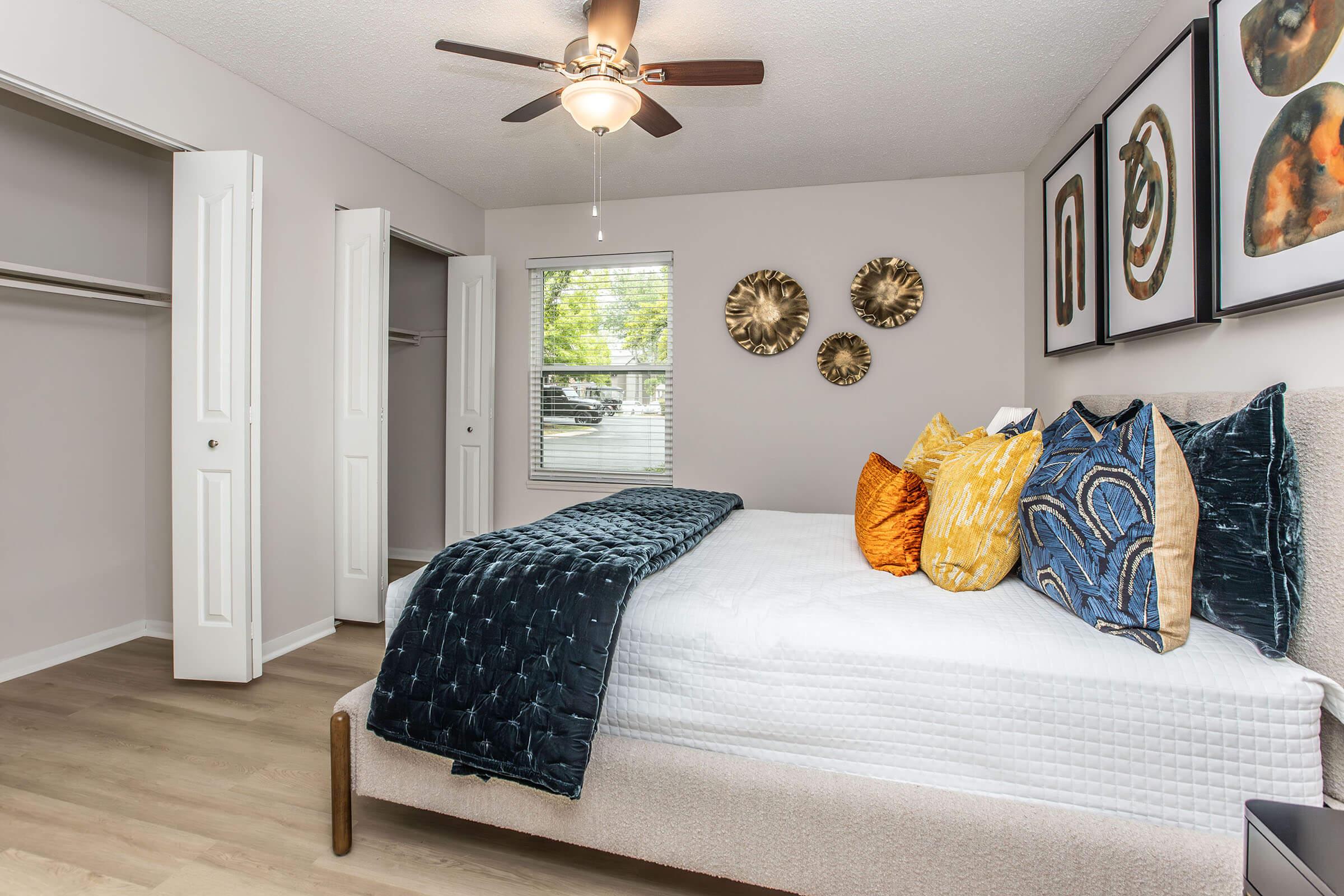 A well-decorated bedroom featuring a queen-sized bed with blue and orange decorative pillows, a cozy quilt, and a ceiling fan. The room includes two closet doors and framed artwork on the walls, contributing to a modern and inviting atmosphere. Natural light enters through a window.