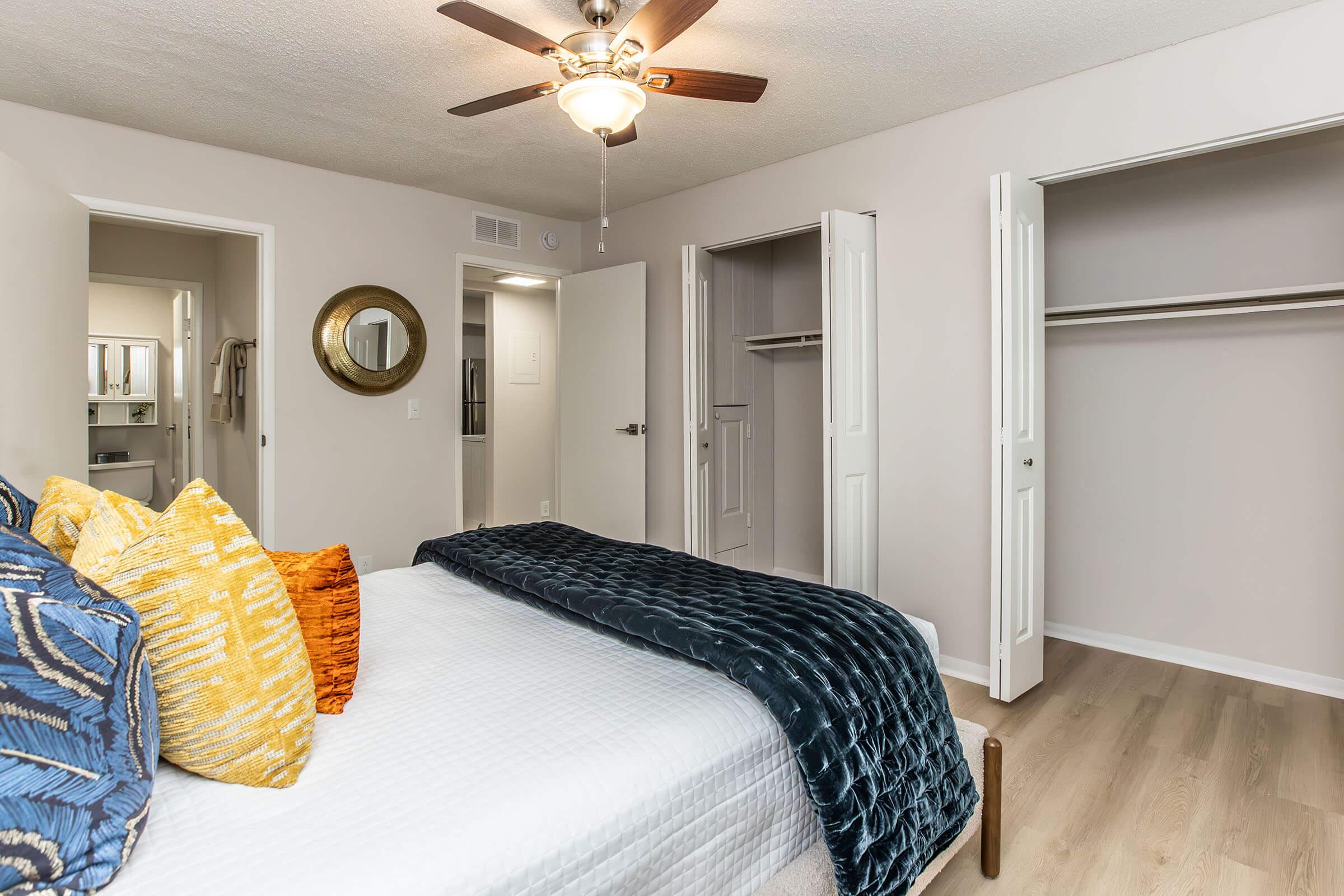 A well-lit bedroom featuring a queen-sized bed with colorful throw pillows and a textured blanket. The room has light-colored walls, a ceiling fan, and wood flooring. There are open closets with sliding doors and a mirror on the wall reflecting the space, creating a cozy and inviting atmosphere.