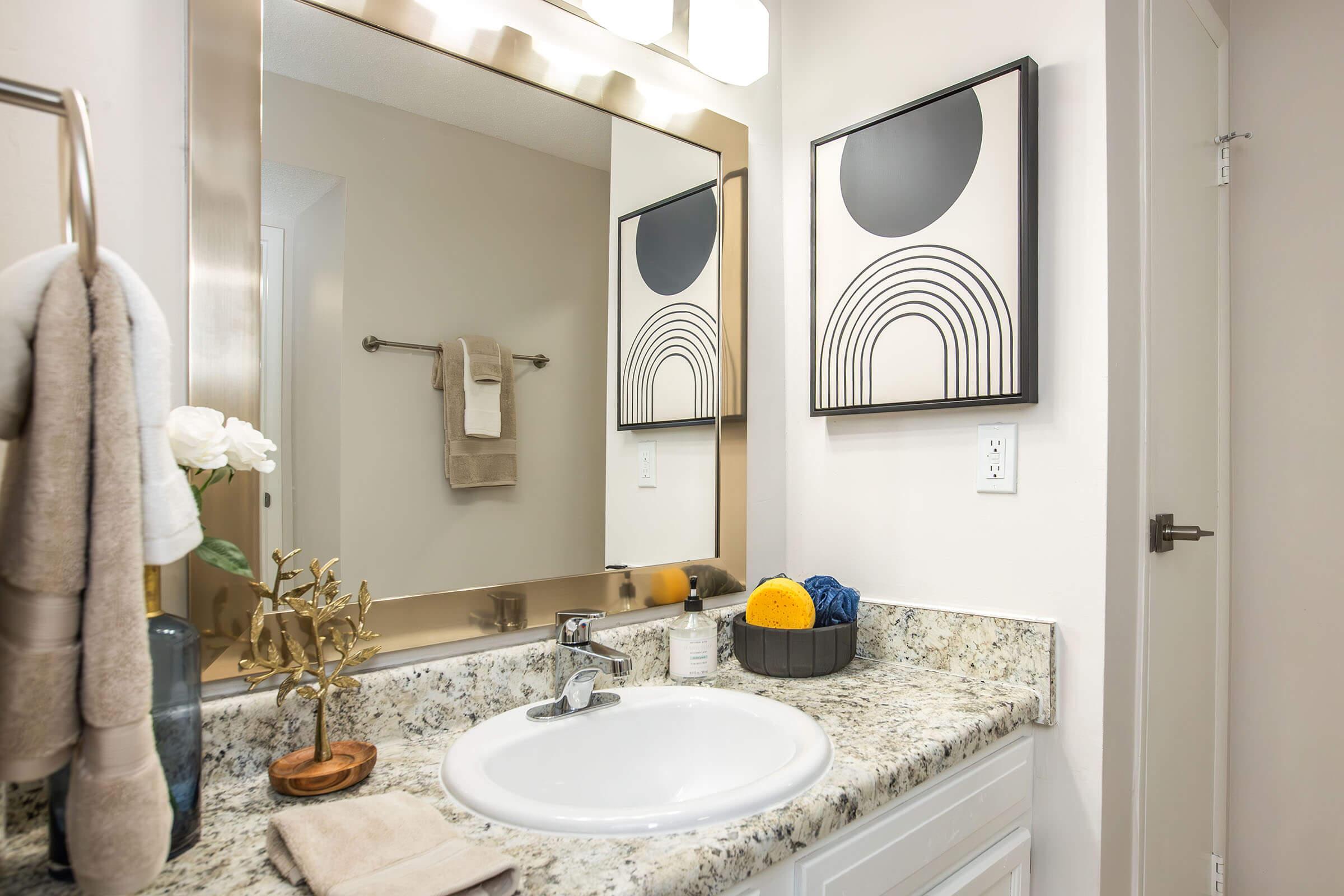 A modern bathroom featuring a stylish sink with a granite countertop. A large mirror above the sink reflects the space, and there are two black and white framed artworks on the wall. A towel rack holds a beige towel, while decorative items like a small plant and colorful sponges are arranged nearby.
