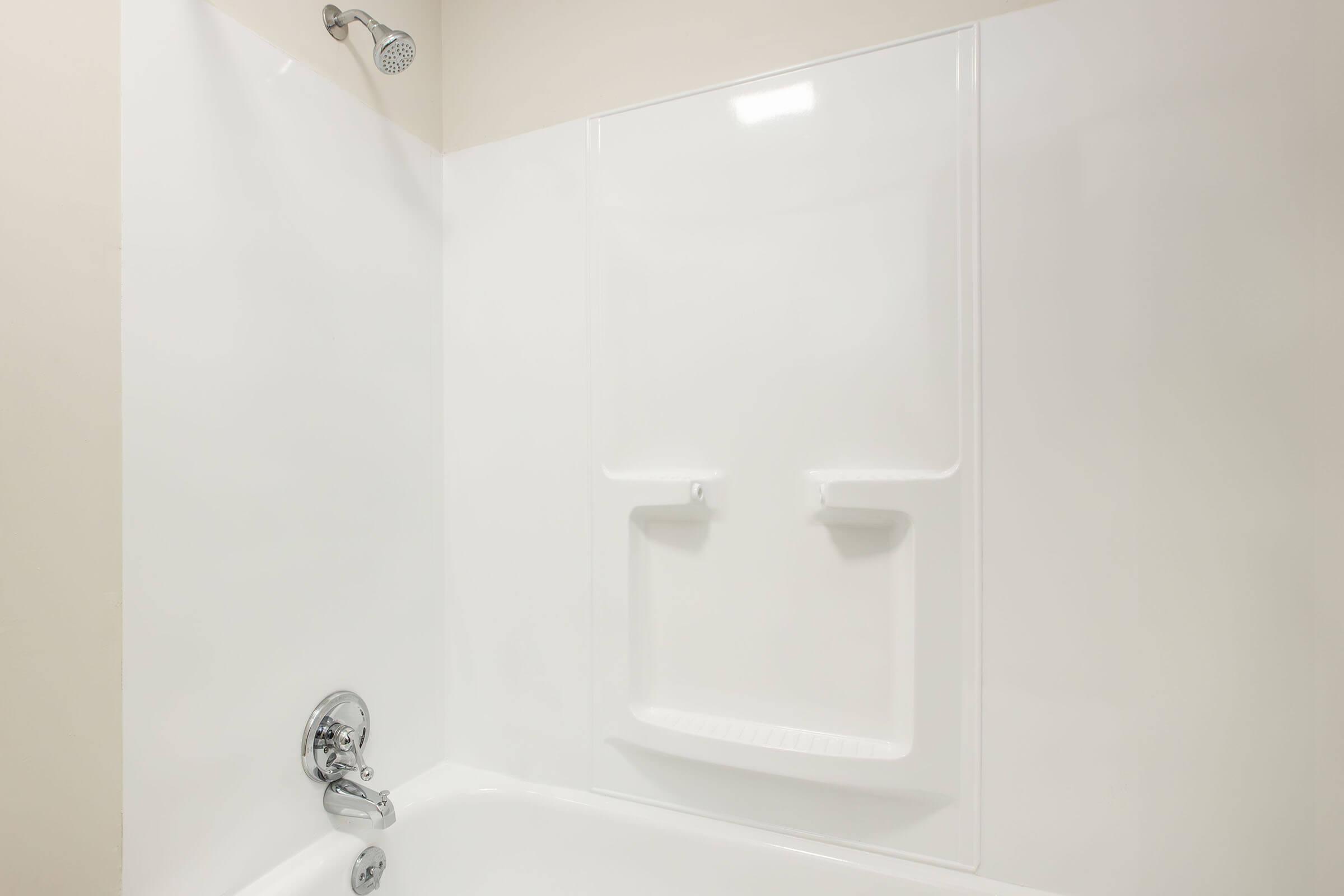 A clean, white bathtub and shower stall with a chrome showerhead. The walls are smooth and glossy, and there are two built-in shelves for toiletries. The overall look is modern and minimalistic, showcasing a bright and airy bathroom space.