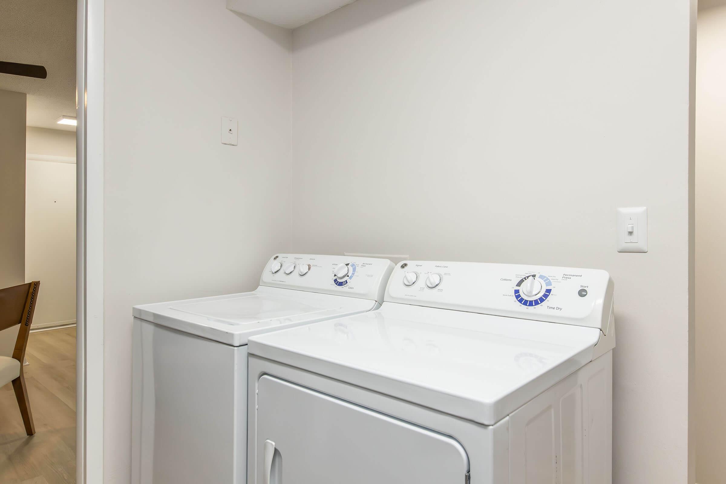 A laundry area featuring a white washing machine and a white dryer side by side, against a plain wall. There is a light-colored floor visible, and a hint of furniture or an interior space in the background, creating a clean and functional laundry space.
