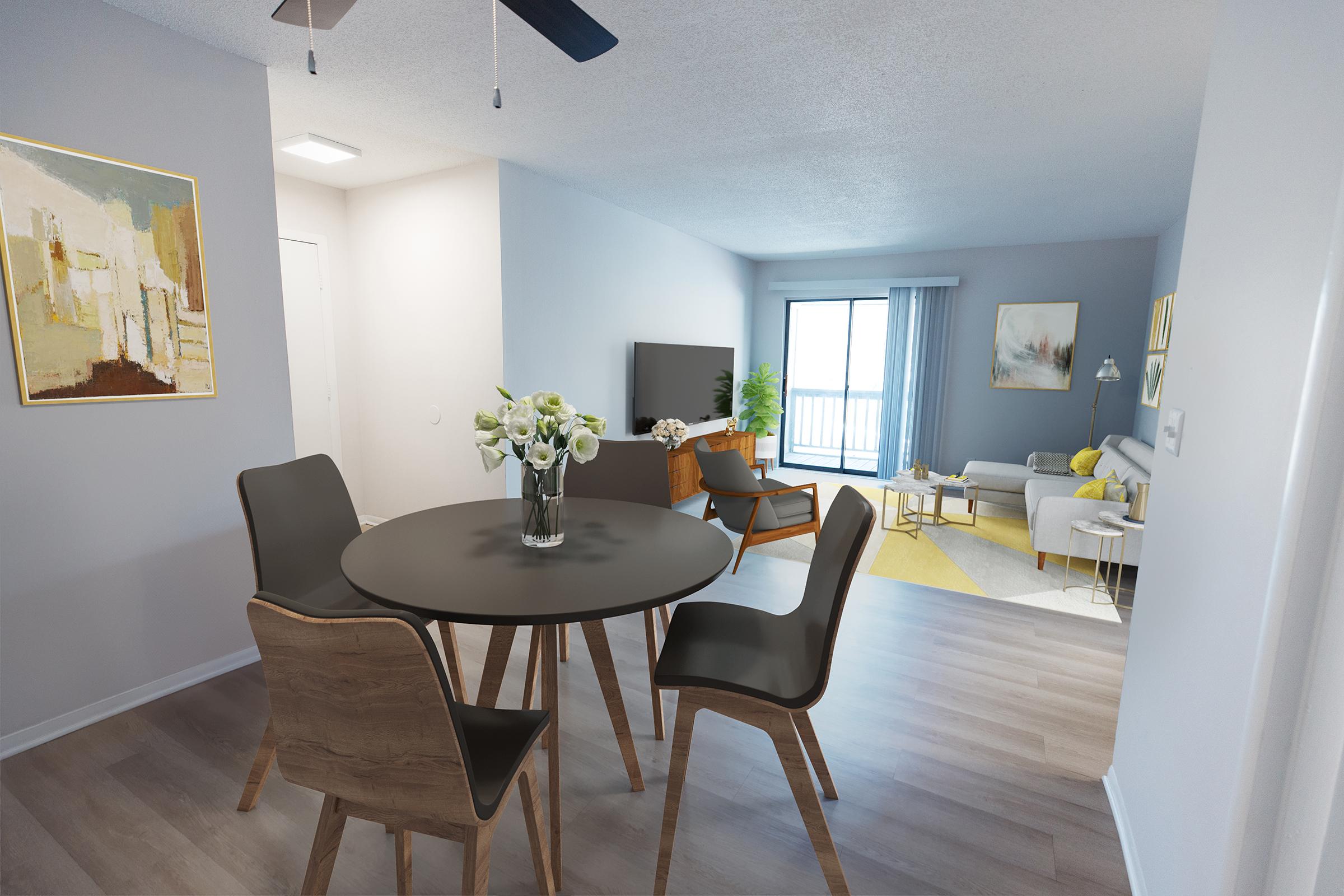 A modern living space featuring a round dining table with four chairs, a vase of flowers, a television, and a comfortable sofa. Natural light enters through a sliding glass door leading to a balcony, with light gray walls and hardwood floors, creating a bright and inviting atmosphere.